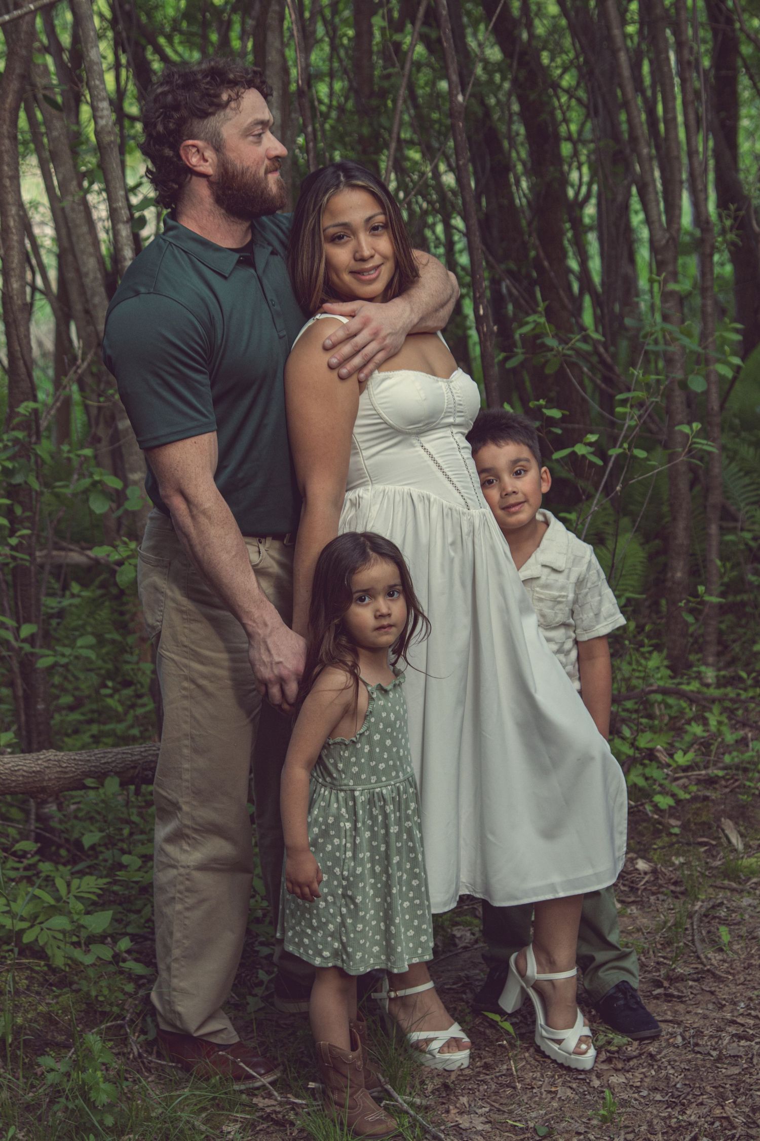 A family poses together in a wooded forest setting showing affection and wearing casual attire.