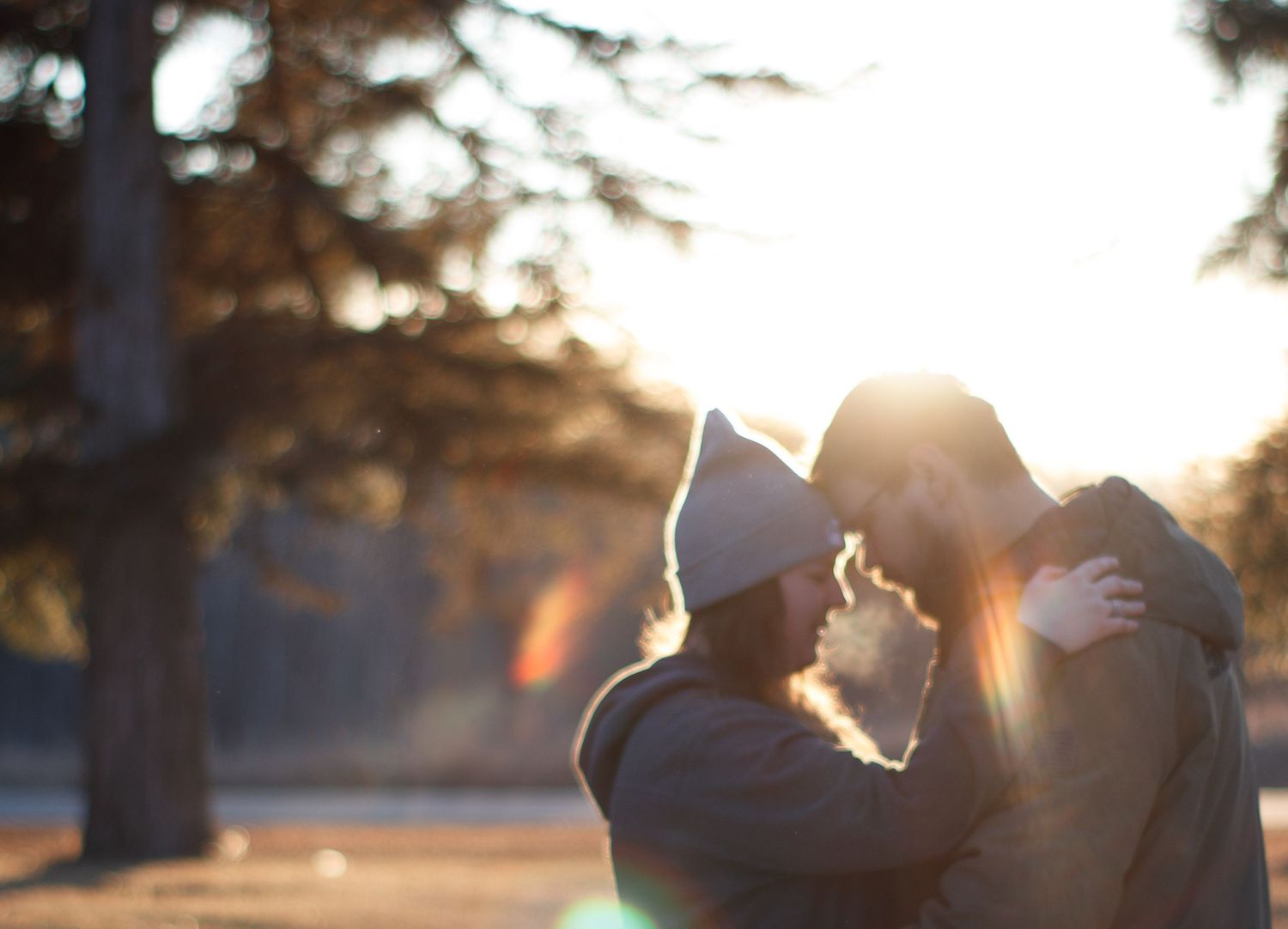Silhouetted figures embrace outdoors at sunset with warm backlit glow and lens flare.