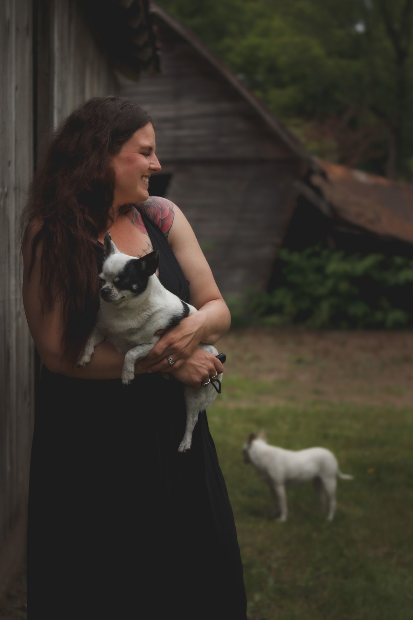 Smiling woman with long hair holds a small black and white dog outdoors near a rustic barn, with a white dog in background.