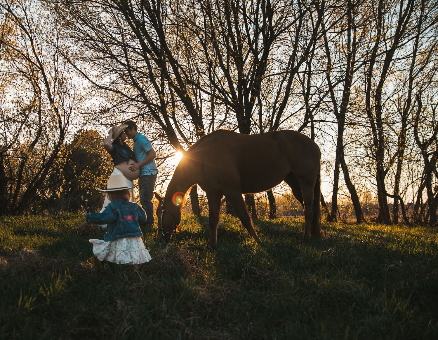 Family with young girl in cowboy hat petting a brown horse at golden sunset among bare trees.