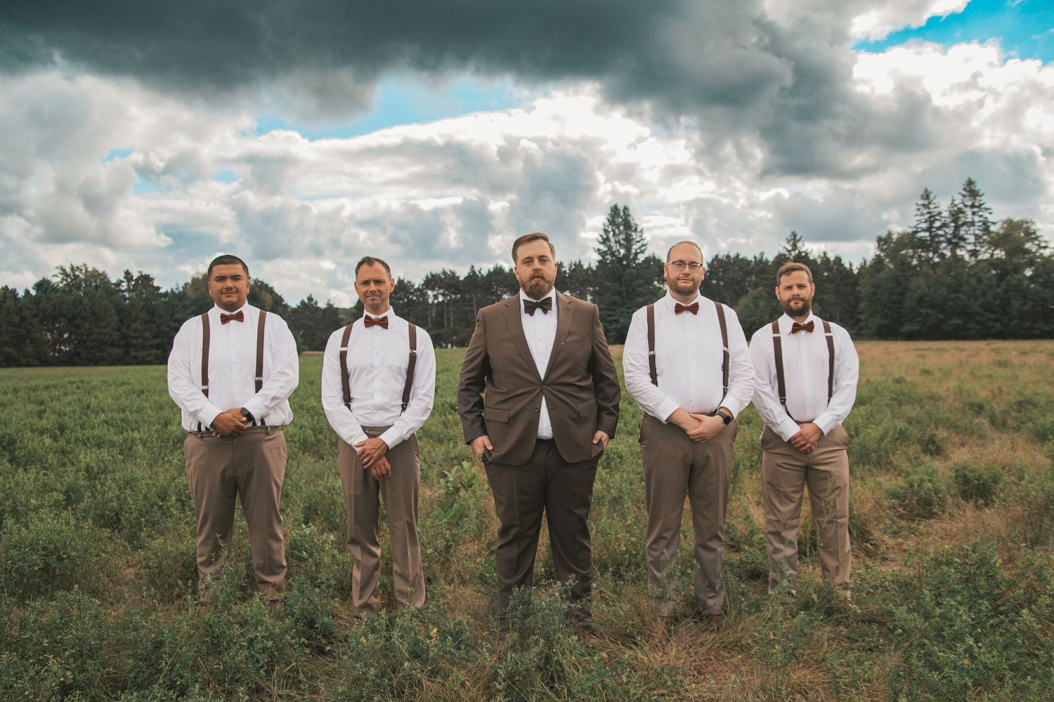 A group of five groomsmen stand in a field wearing suspenders and bowties against dramatic cloudy skies.