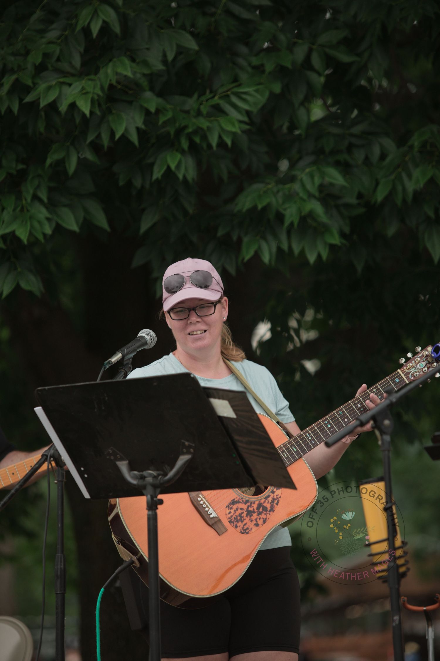 Musicians perform at an outdoor venue with guitars and music stands beneath leafy tree cover.