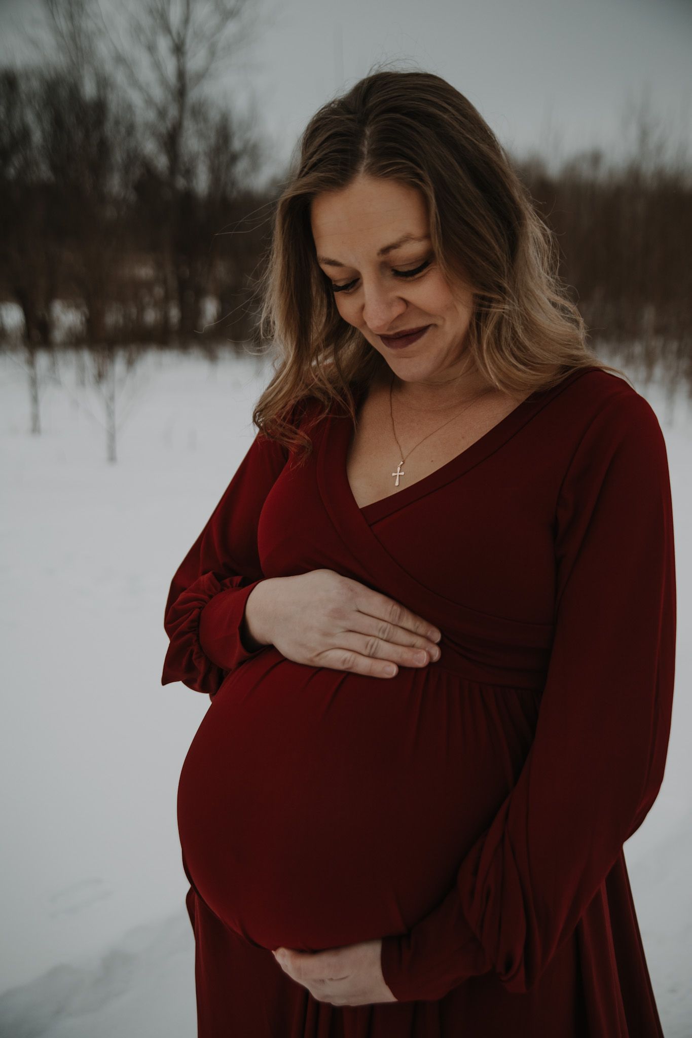 Expectant mother in burgundy dress cradles baby bump against snowy winter backdrop.