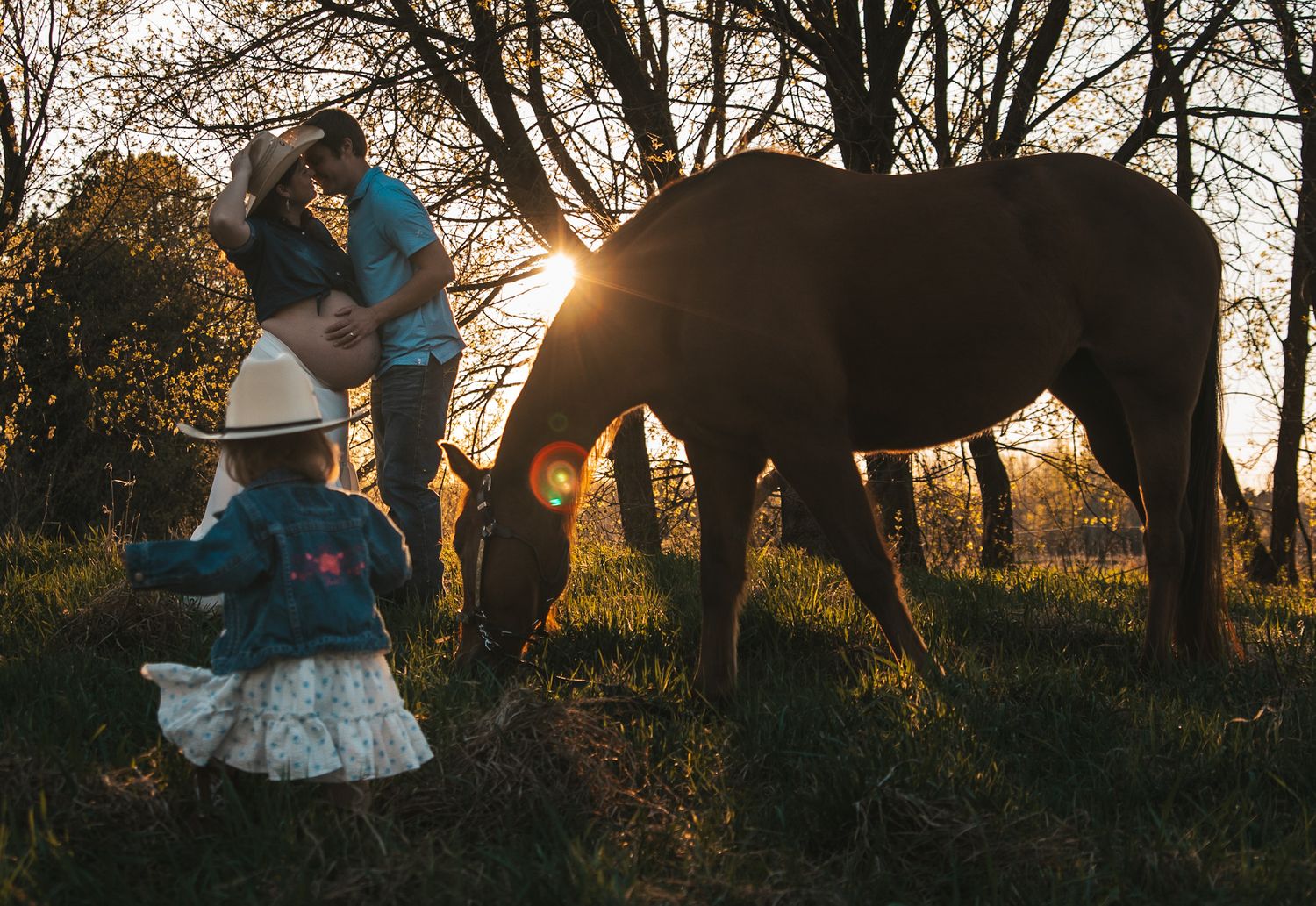 Family with pregnant mother and toddler standing beside a grazing brown horse at golden sunset among trees.