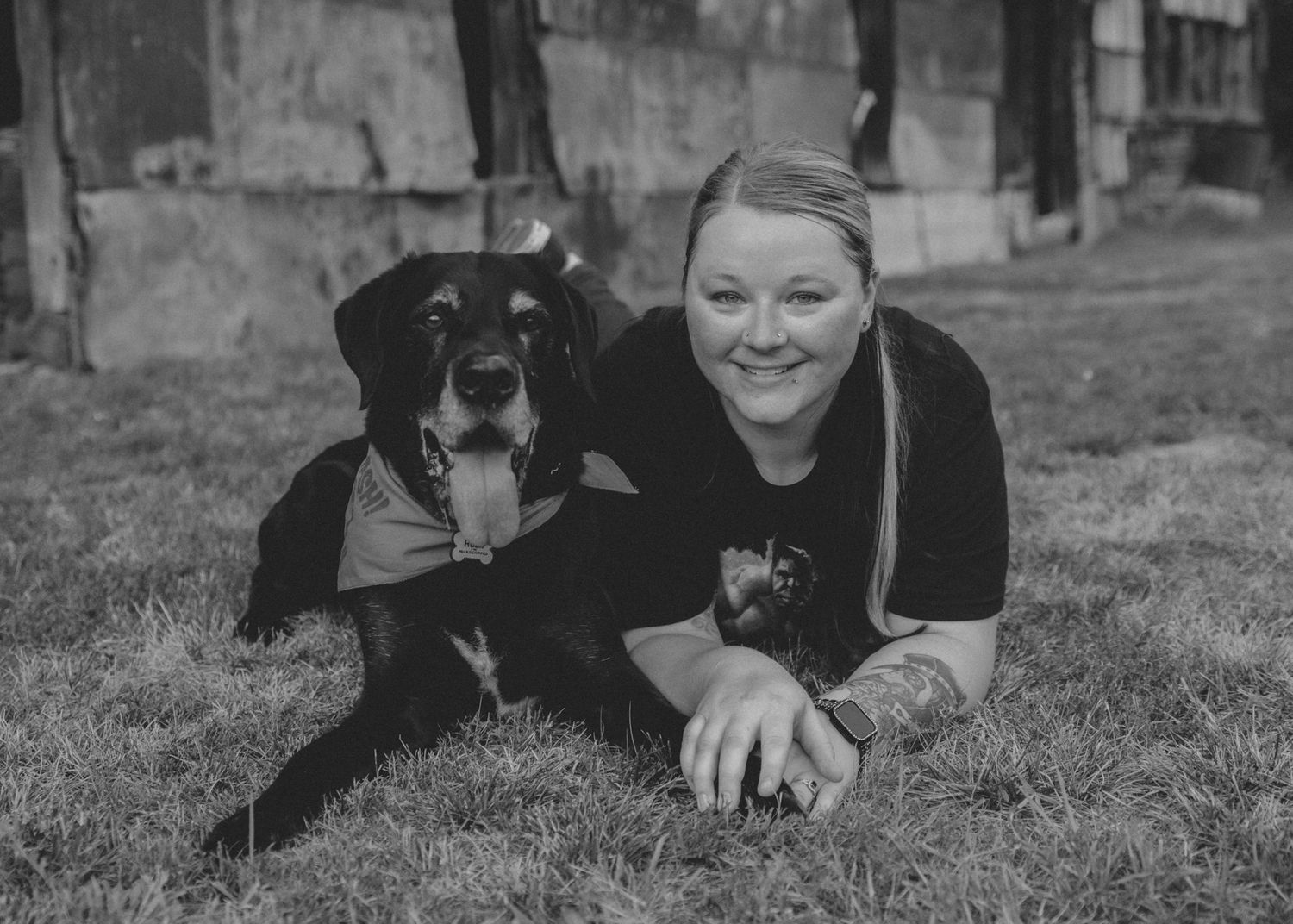 Woman smiling while lying on grass next to her black dog in a black and white photo.