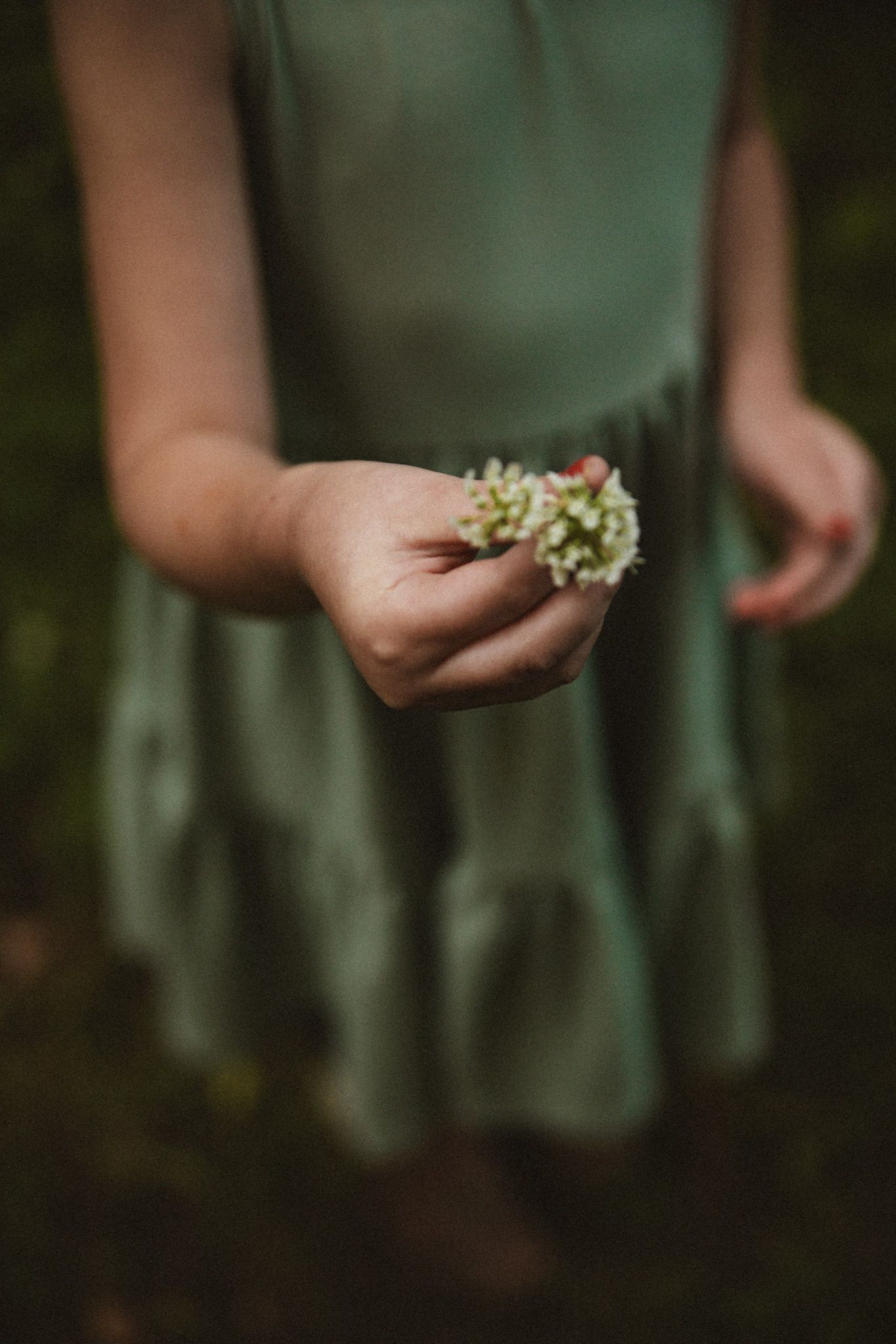 A young girl in a green dress holds a small white clover flower in her outstretched hand outdoors.