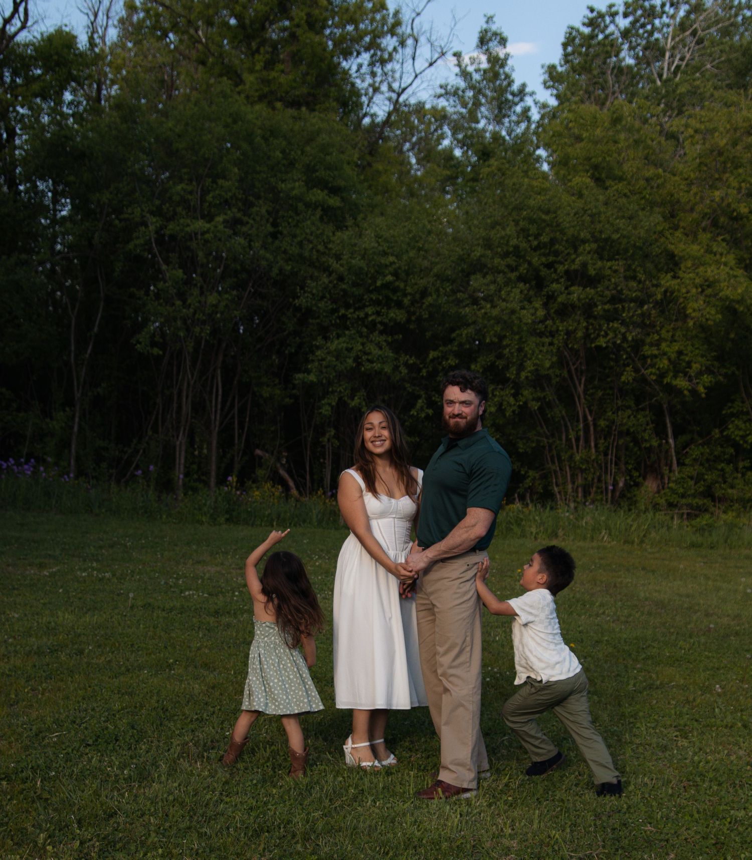 A family poses playfully together on a grassy field with trees in the background at dusk.