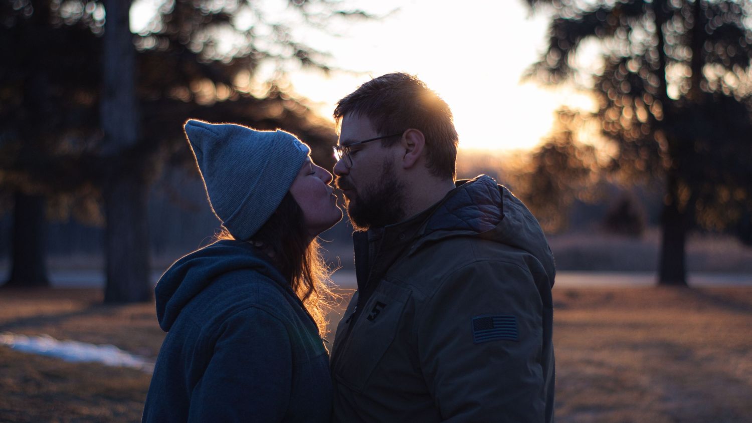 A silhouette of a couple shares a tender embrace in front of a glowing sunset through trees.