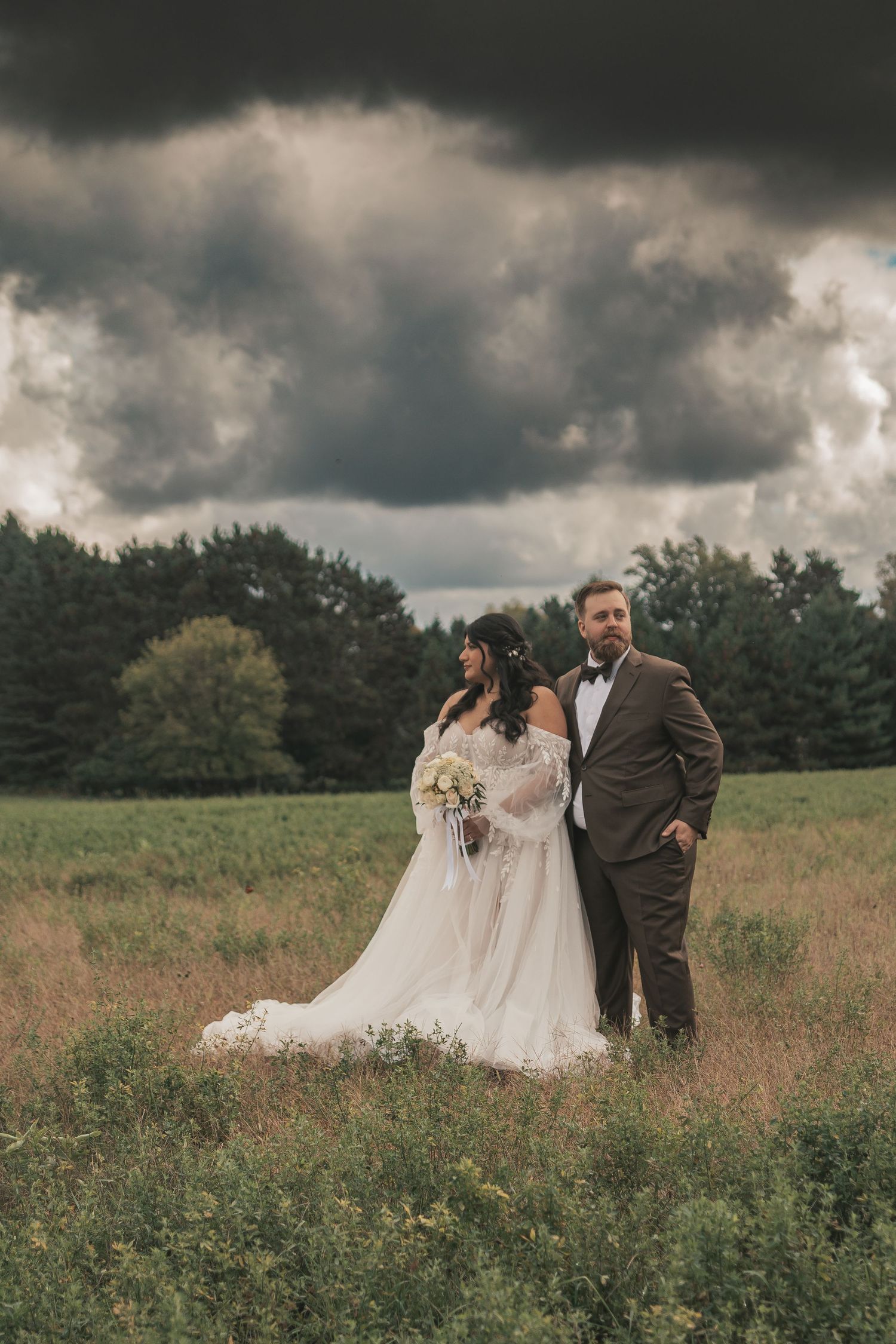 A couple poses together in a meadow with dramatic storm clouds overhead during their wedding photoshoot.