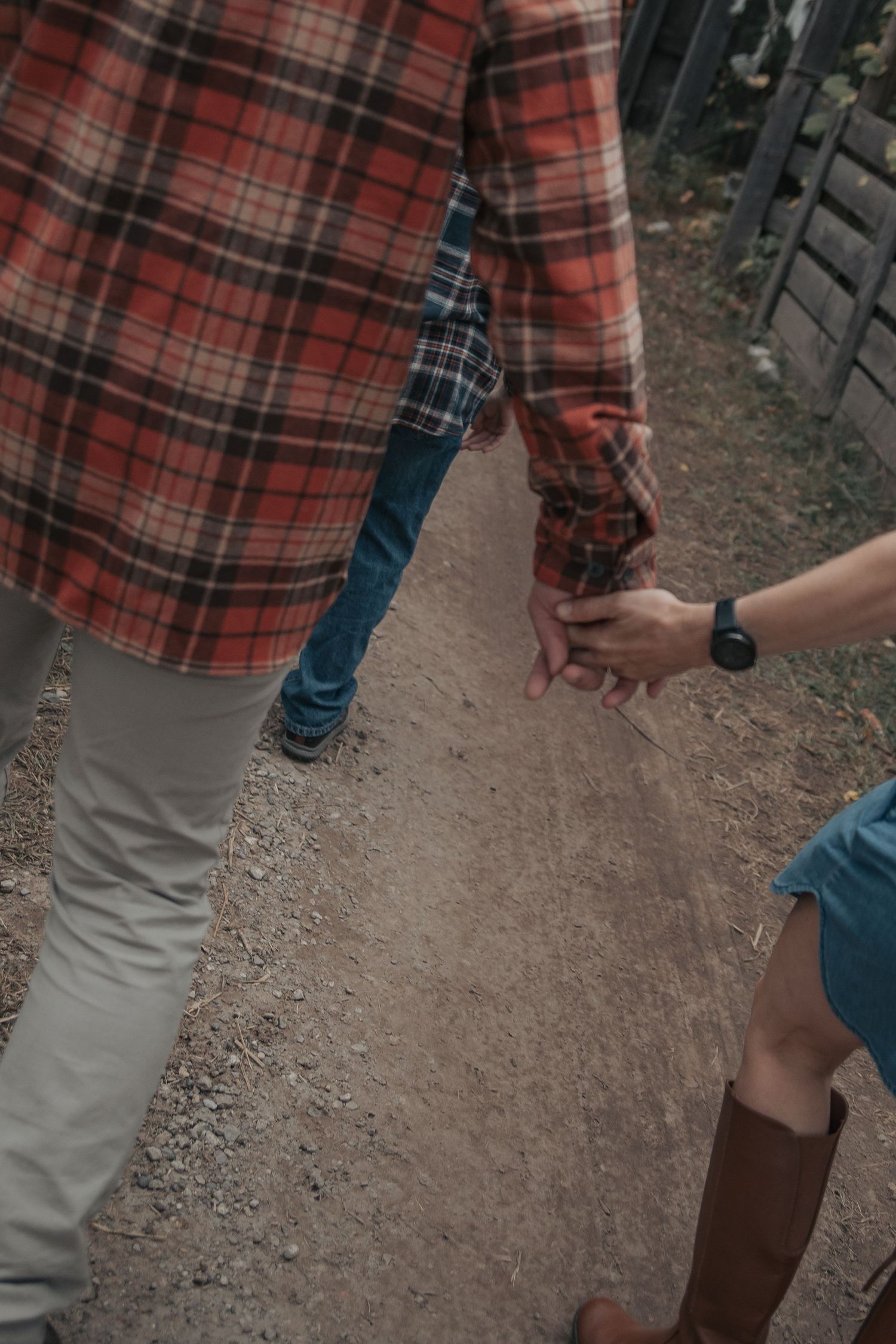 Two people holding hands while standing on a dirt path, one wearing a red plaid flannel shirt and khaki pants.