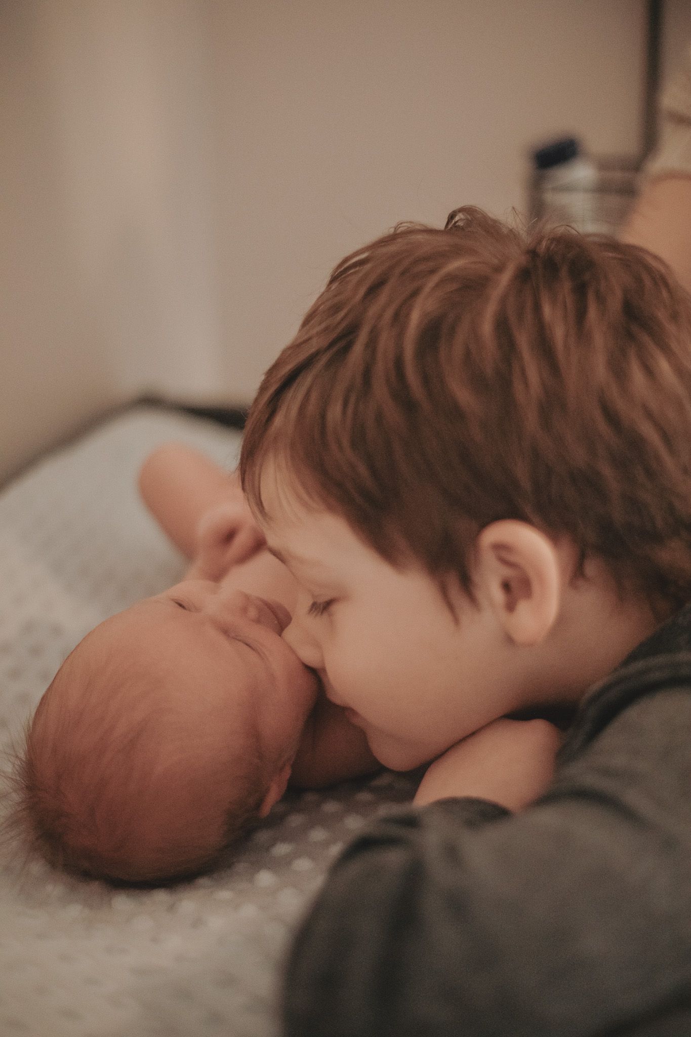 Young boy gently kissing a sleeping newborn baby sibling on a soft blanket in a tender moment.