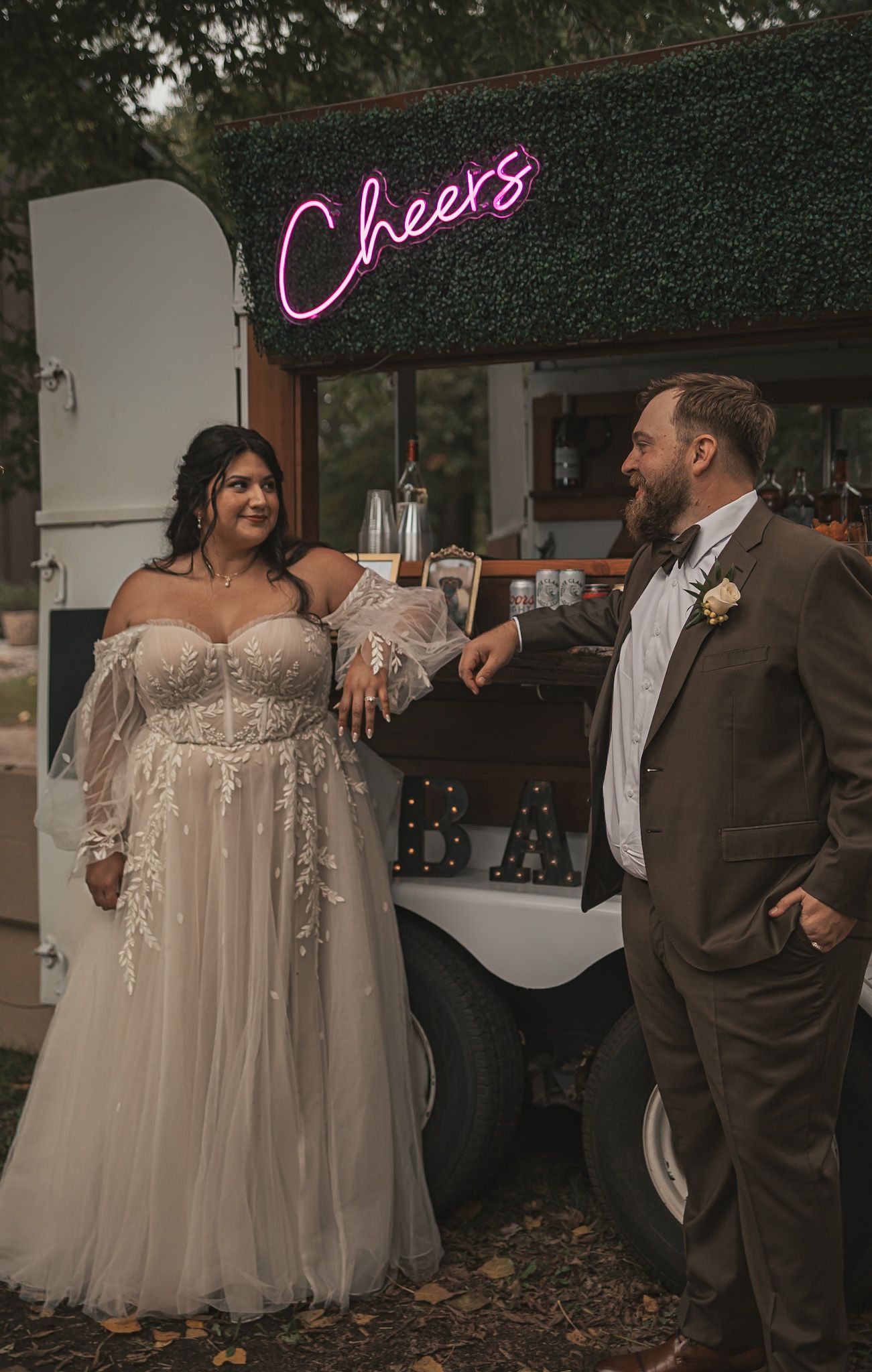 A couple shares a celebratory toast at their wedding reception under a neon 'Cheers' sign against a greenery backdrop.