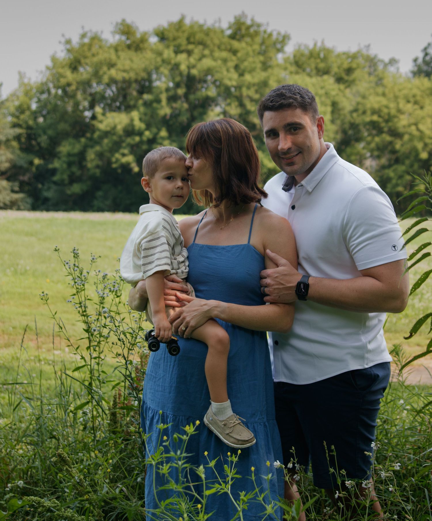 A sweet outdoor family portrait captured in a meadow with wildflowers and trees in the background.