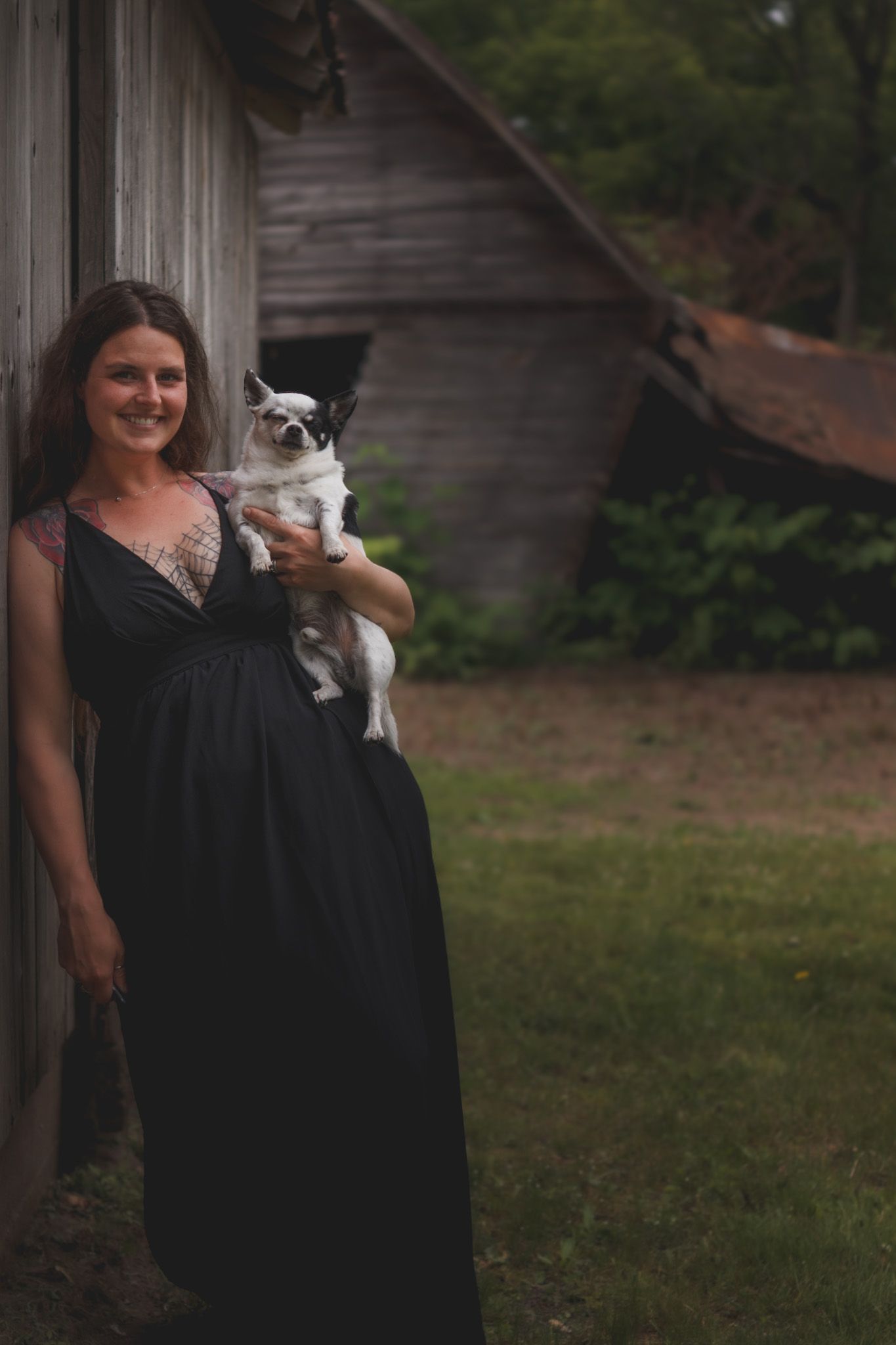 Pregnant woman in black maxi dress holds small chihuahua dog while leaning against rustic barn outdoors.
