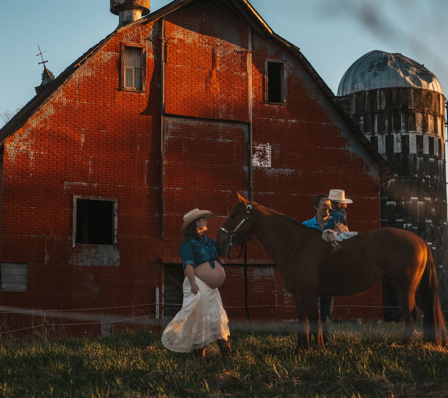Pregnant woman in cowboy hat leads horse with child rider past a red brick barn and grain silo.