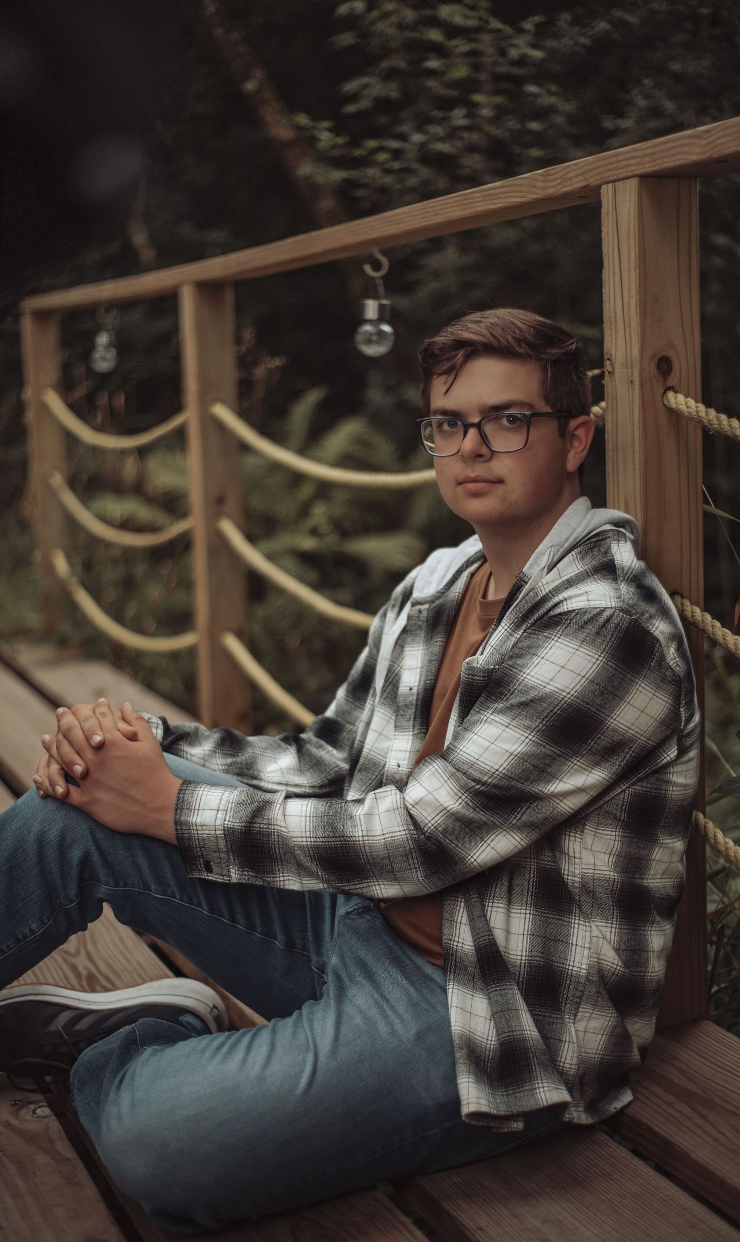 Young man in plaid shirt and glasses sitting on wooden bridge with rope railing surrounded by greenery.