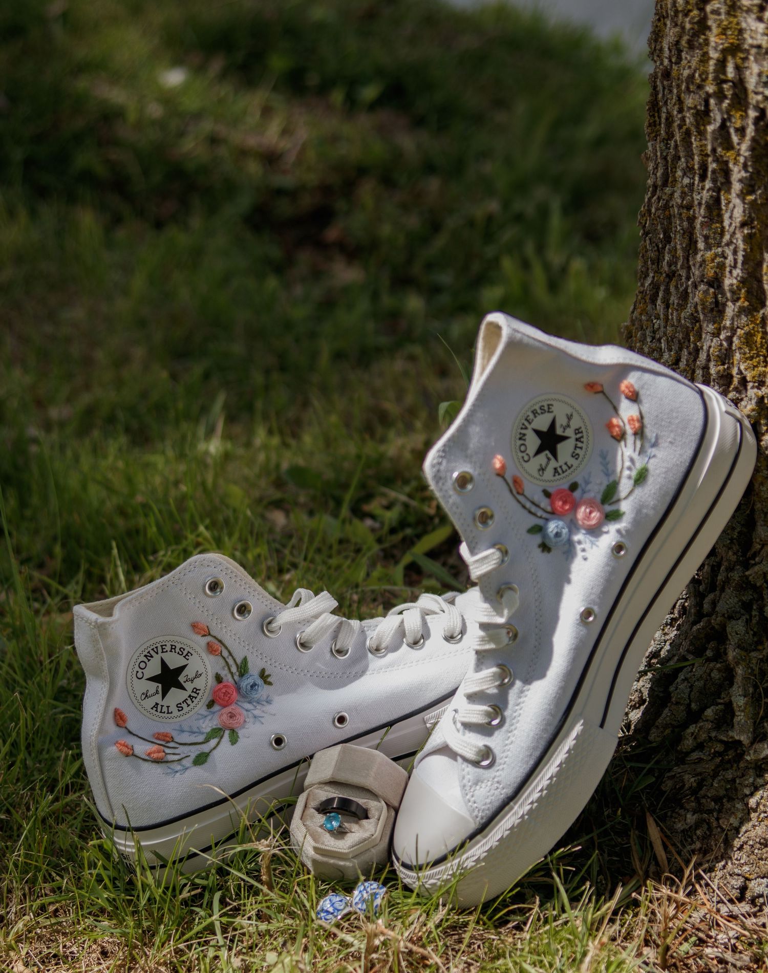 White Converse high-top sneakers decorated with delicate floral designs leaning against a tree trunk.