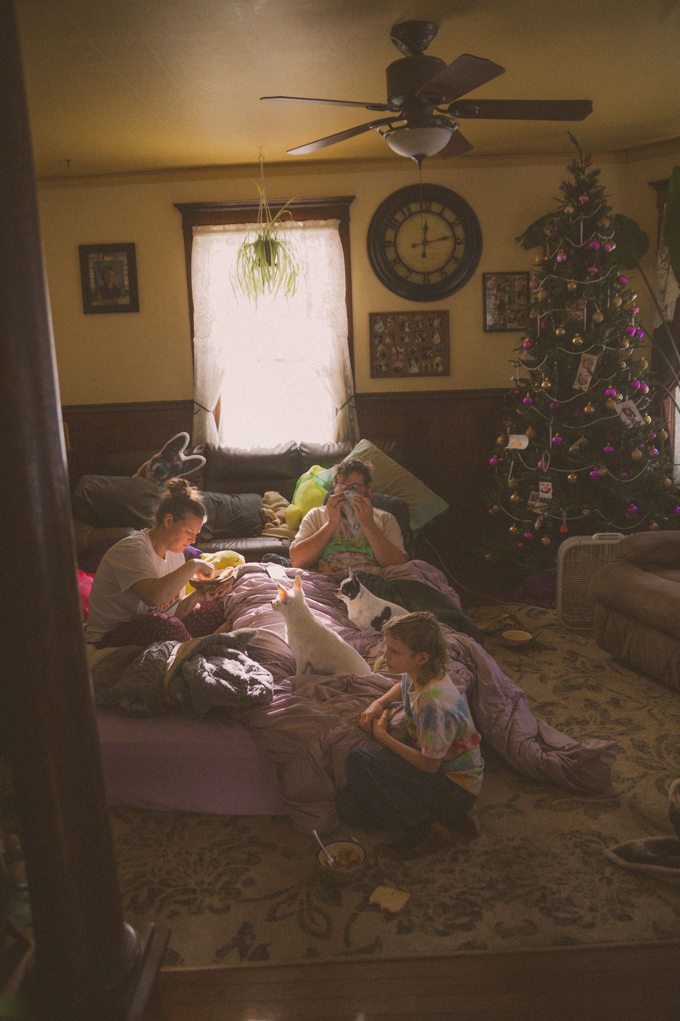 Family relaxing in living room with decorated Christmas tree, ceiling fan, and cozy blankets during holiday season.