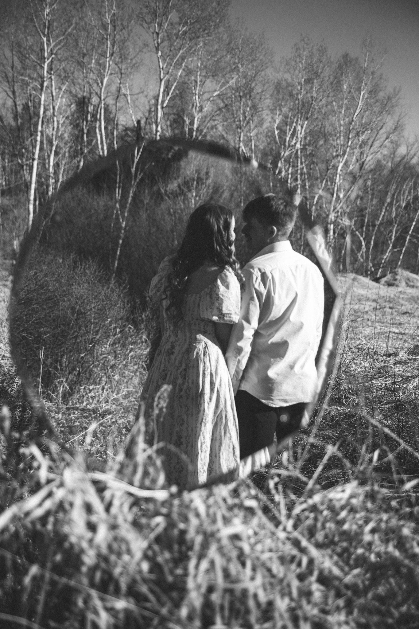 Black and white photo of a romantic couple embracing near birch trees in a field, viewed through a circular frame.