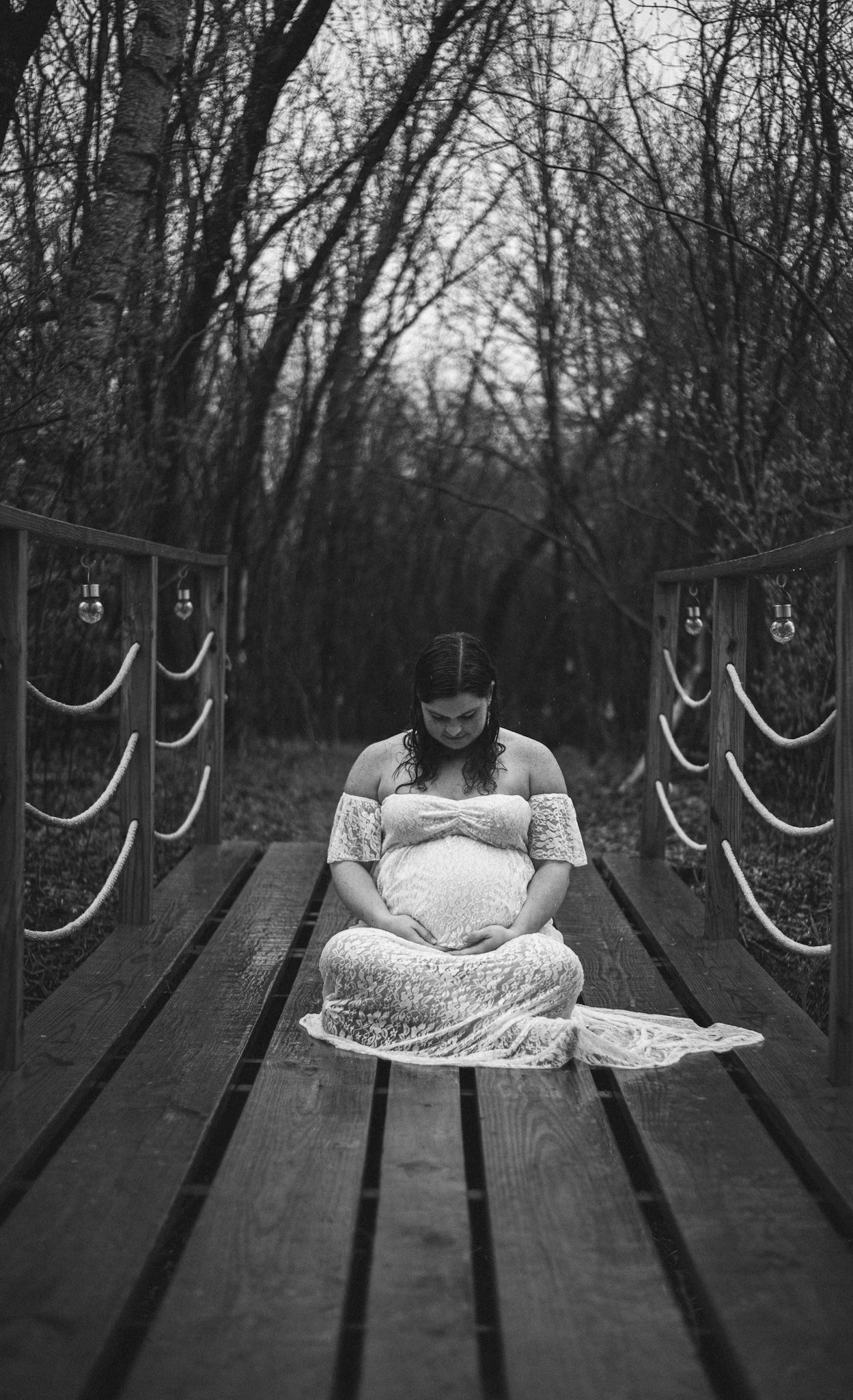 Black and white maternity photo of pregnant woman in lace dress sitting peacefully on a rustic wooden bridge.