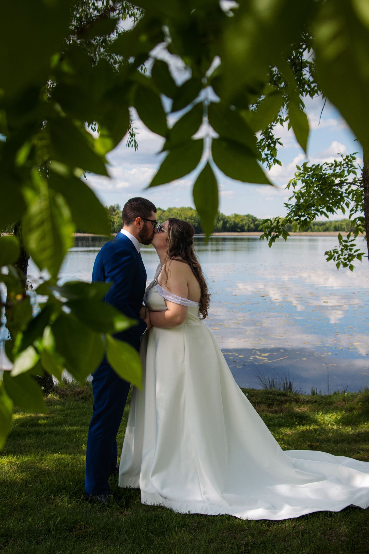 A couple shares a romantic moment by a lake, framed by green leaves with a flowing white wedding dress and blue suit.