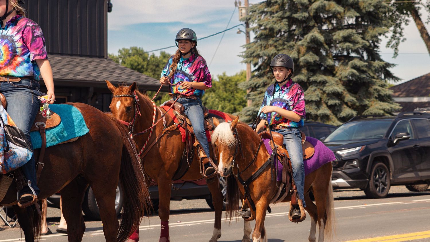 Riders on horseback in western attire form a line along a road during what appears to be a community event.