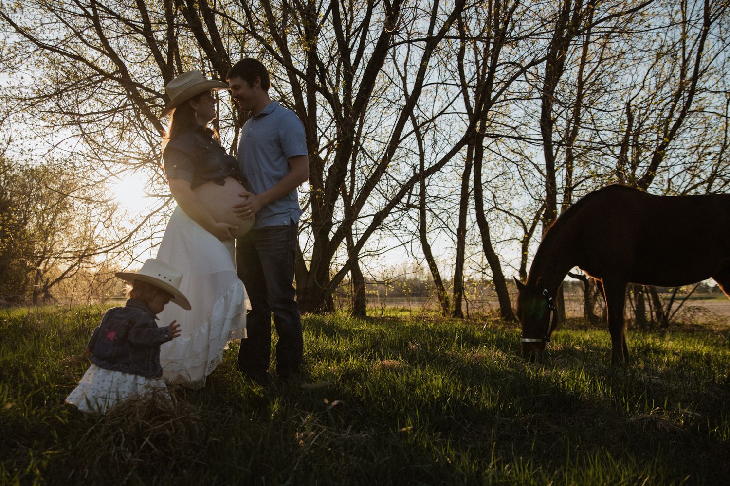 Couple in cowboy hats embrace at golden hour near trees with a grazing horse and child nearby.