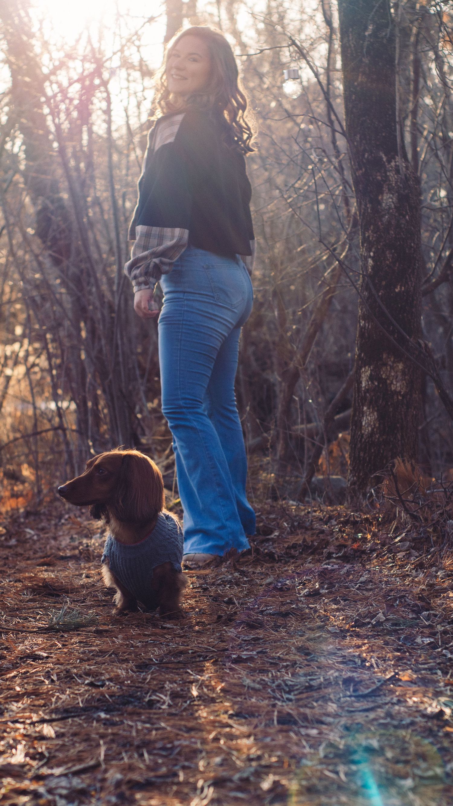 Woman in flare jeans walking with a dachshund on a sunlit forest trail in autumn.