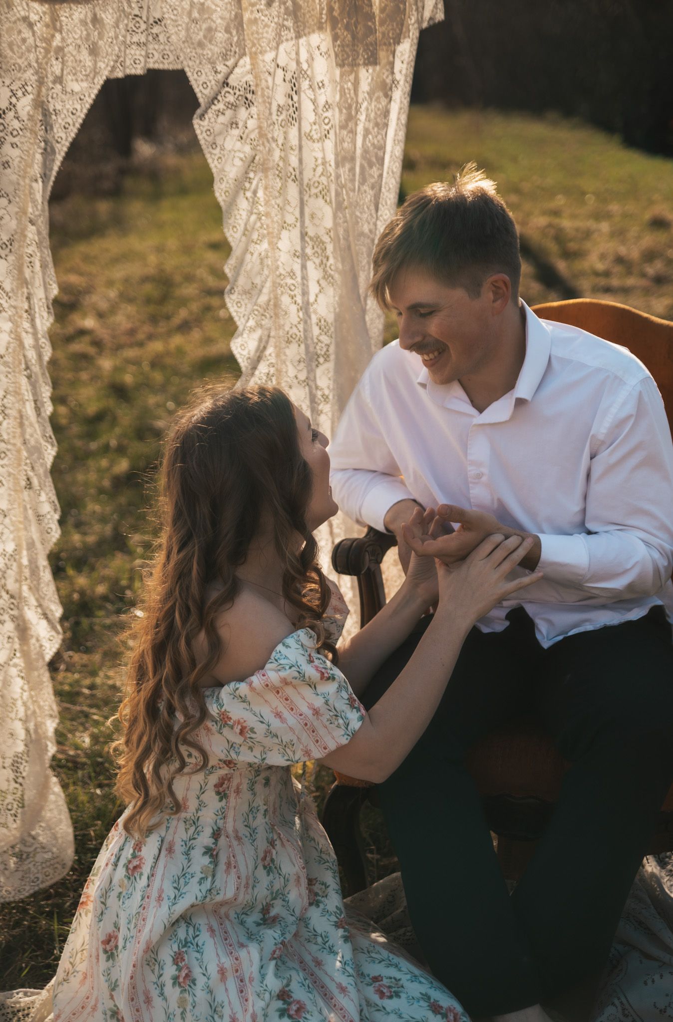 A smiling couple holds hands outdoors near lace curtains, the woman in a floral dress and man in a white shirt.