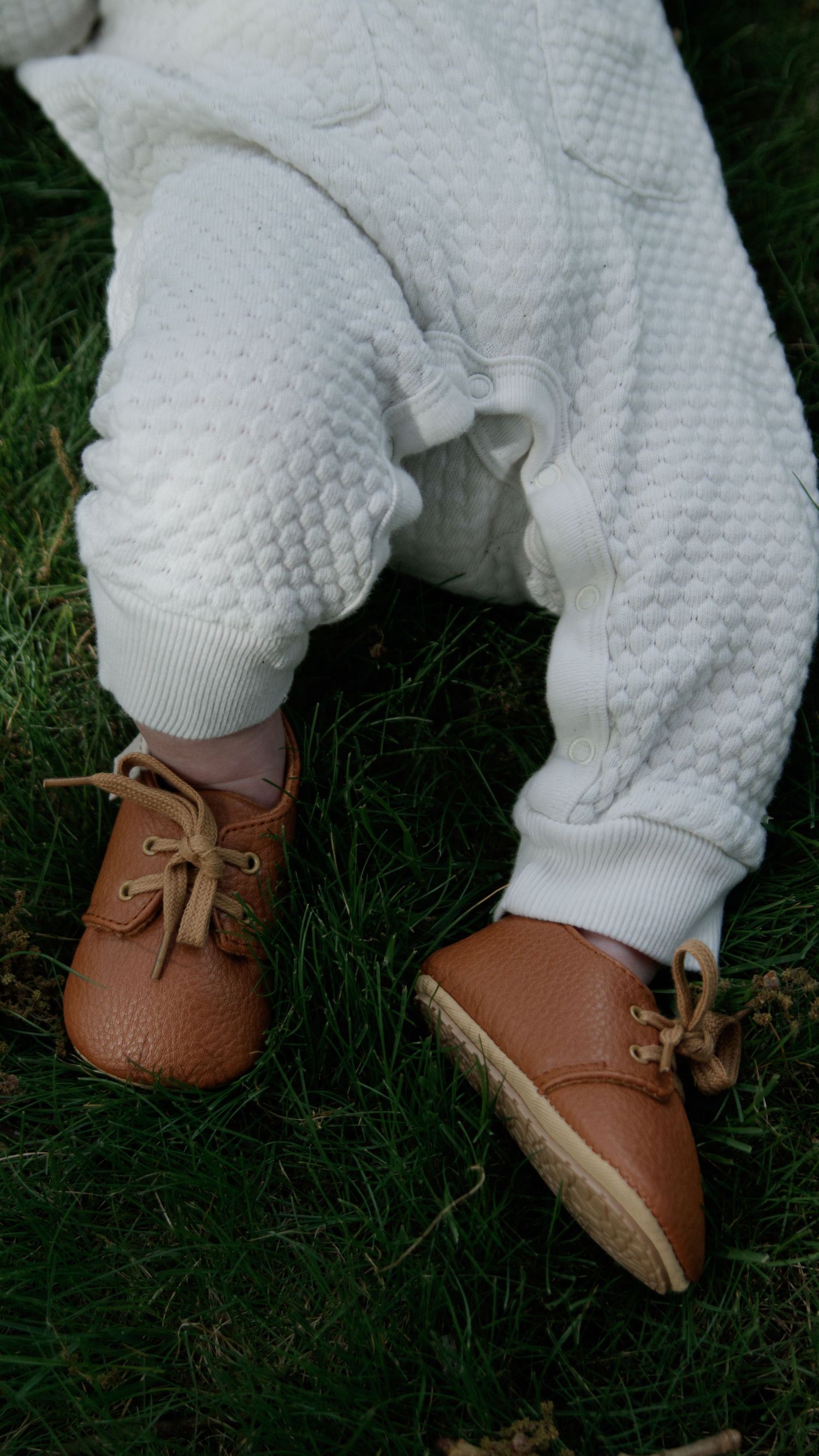 Tiny brown leather baby shoes with white knit socks shown against dark grass in a series of close-up shots.