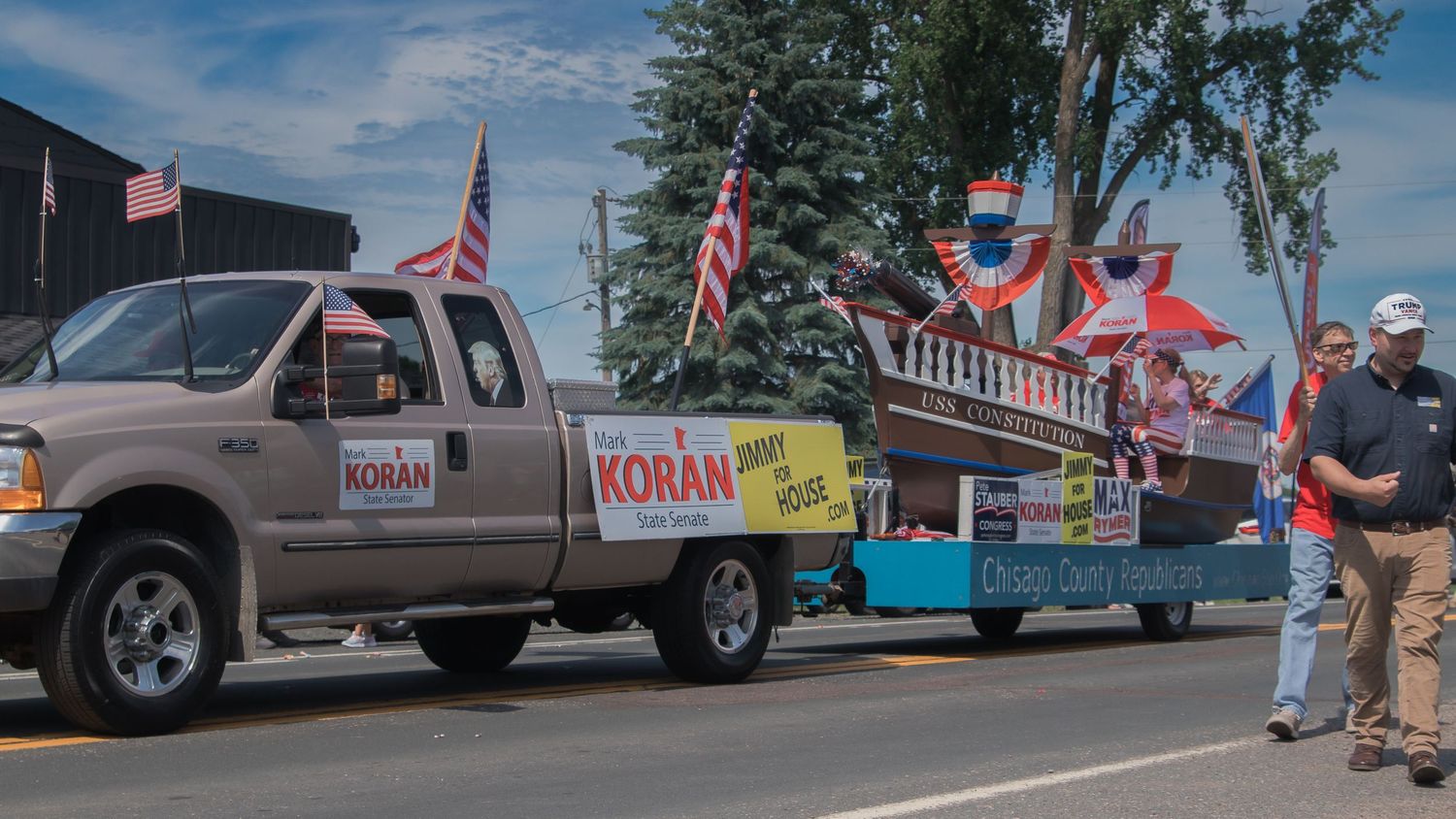 Military and civilian vehicles decorated with flags participate in a community parade on a tree-lined street.