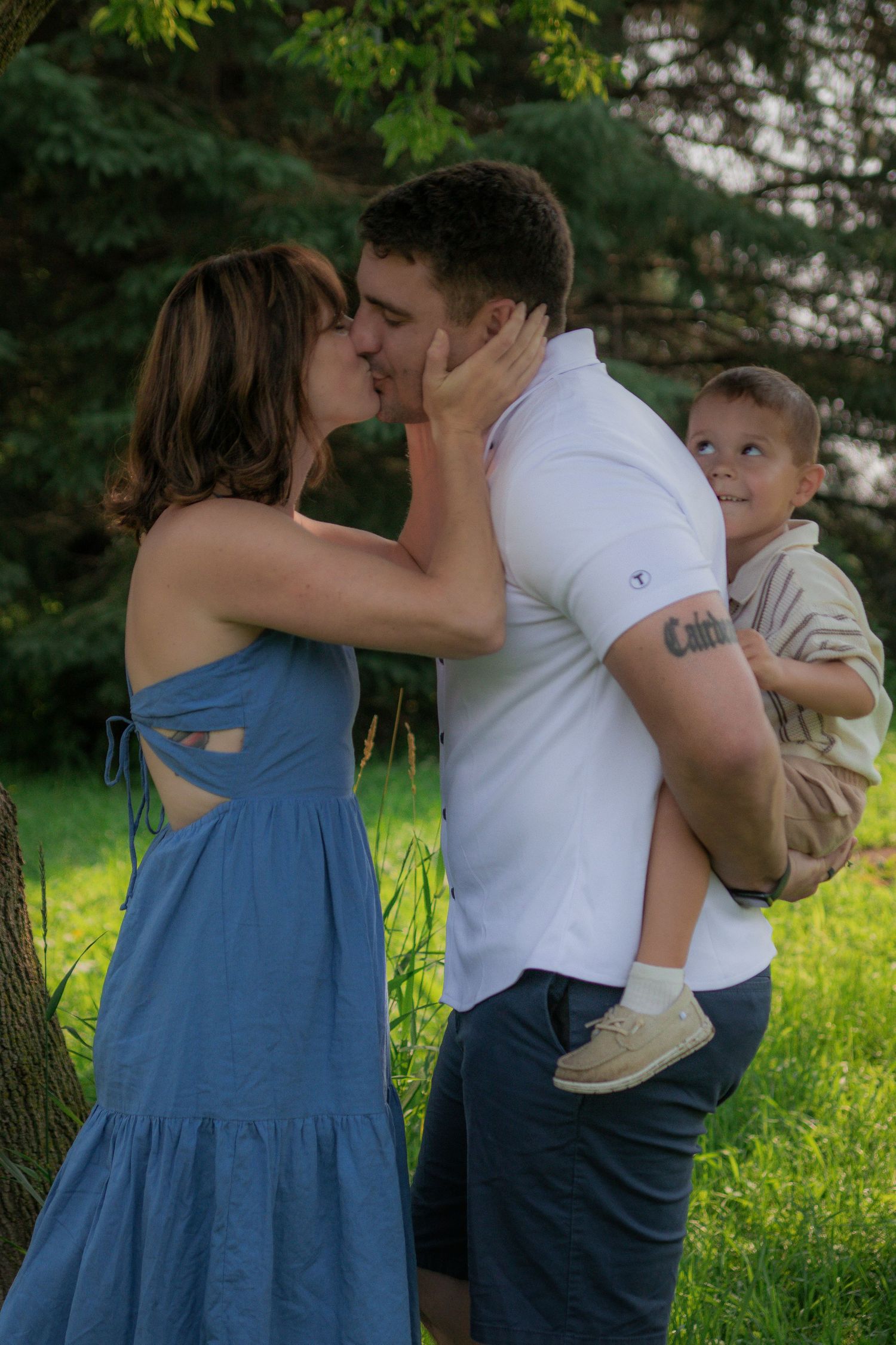 A couple shares a romantic kiss in an outdoor setting while holding a young child on a sunny summer day.