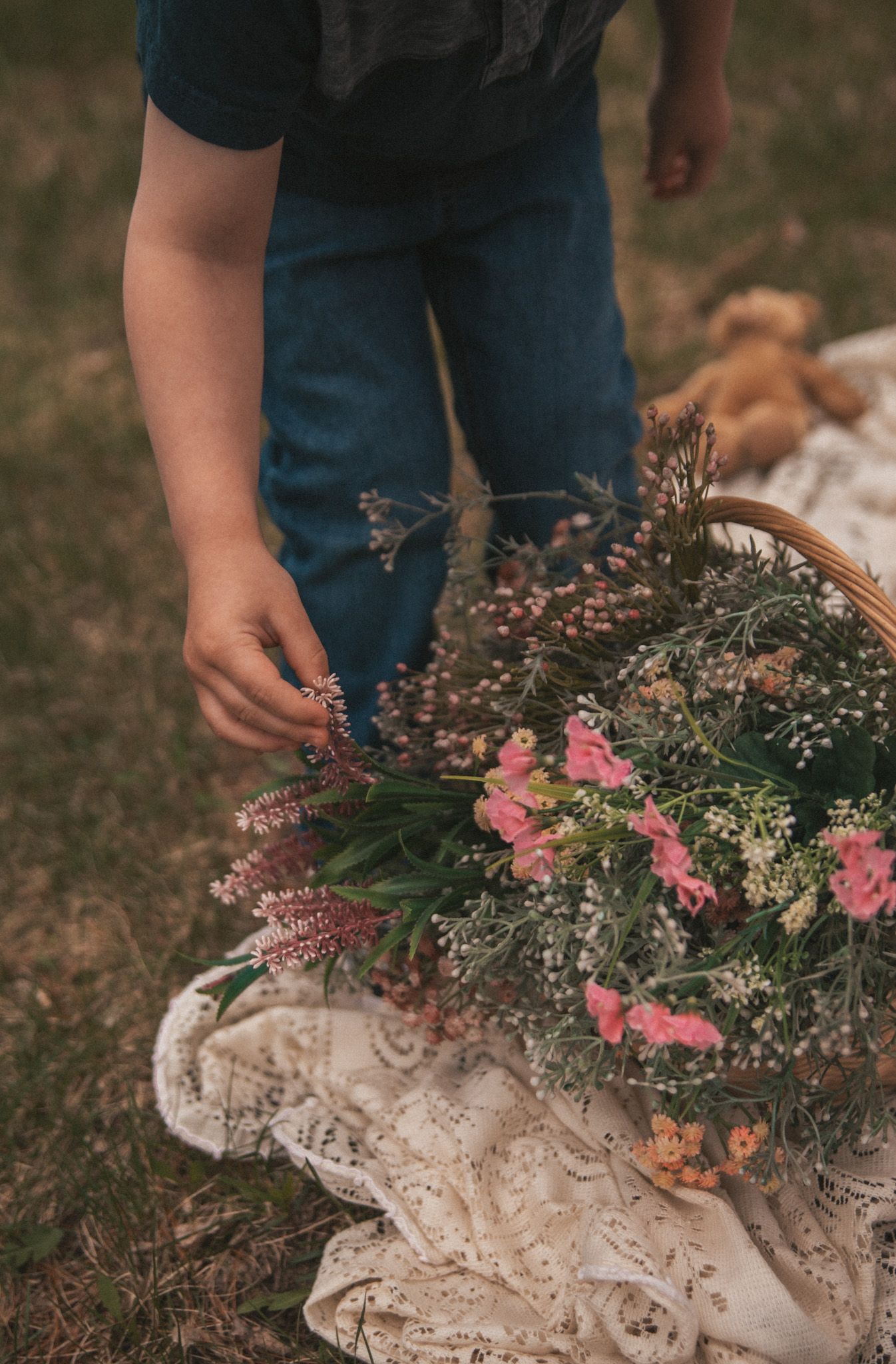 Child arranging pink wildflowers in a wicker basket on a lace blanket outdoors with a teddy bear nearby.