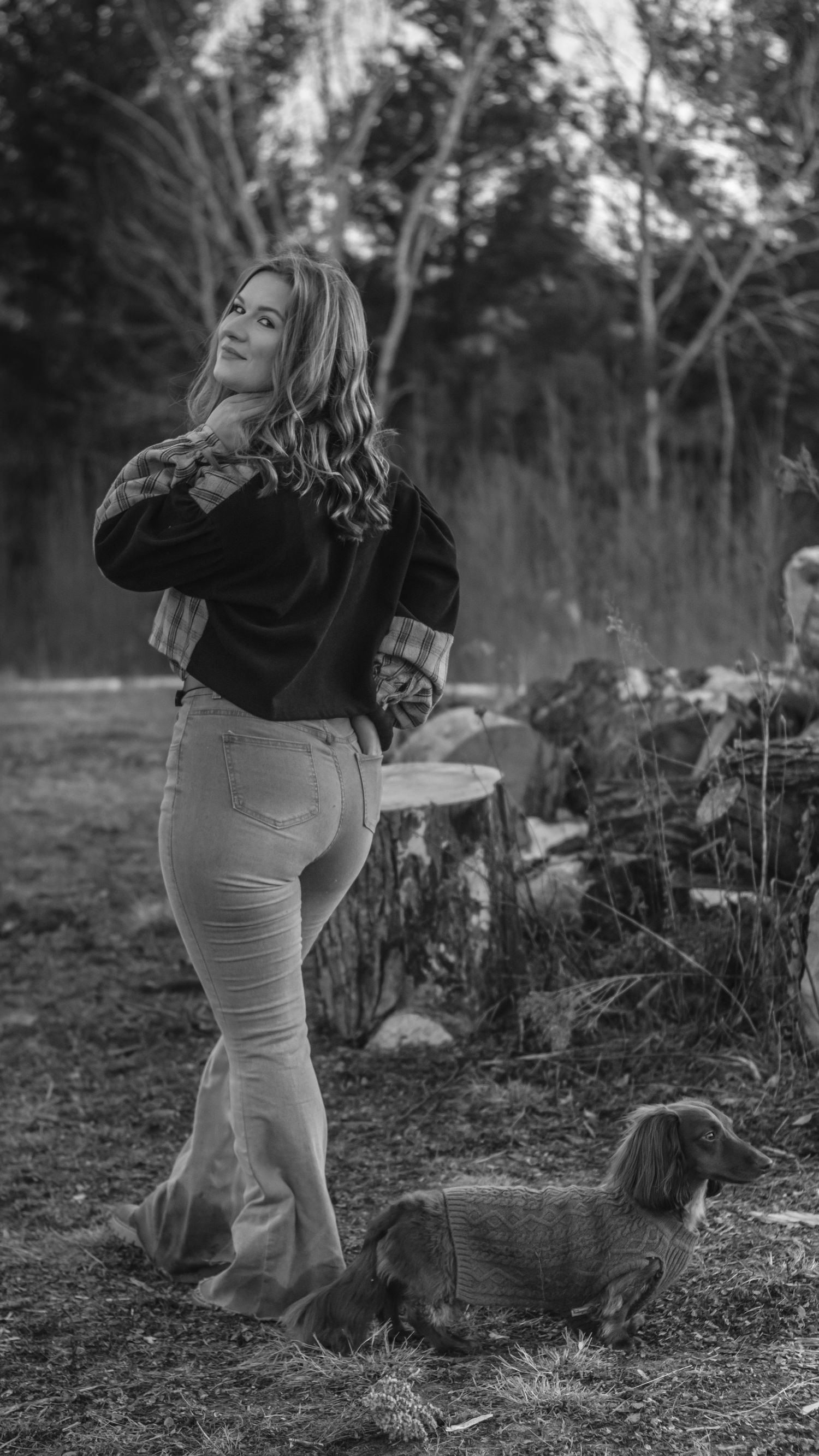 Woman in flare jeans and plaid jacket poses outdoors with a dachshund in black and white photography.