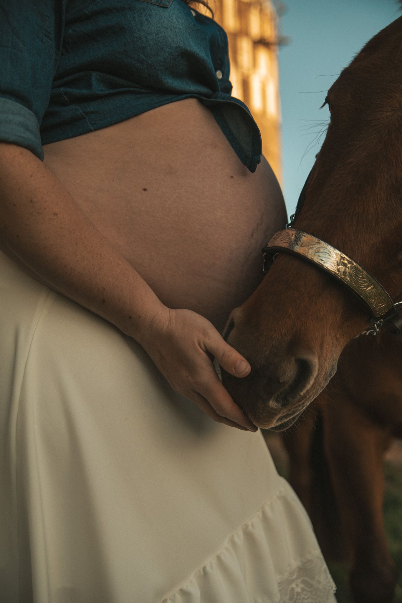 Pregnant woman in white skirt and teal shirt cradling baby bump while a brown horse nuzzles her hand outdoors.