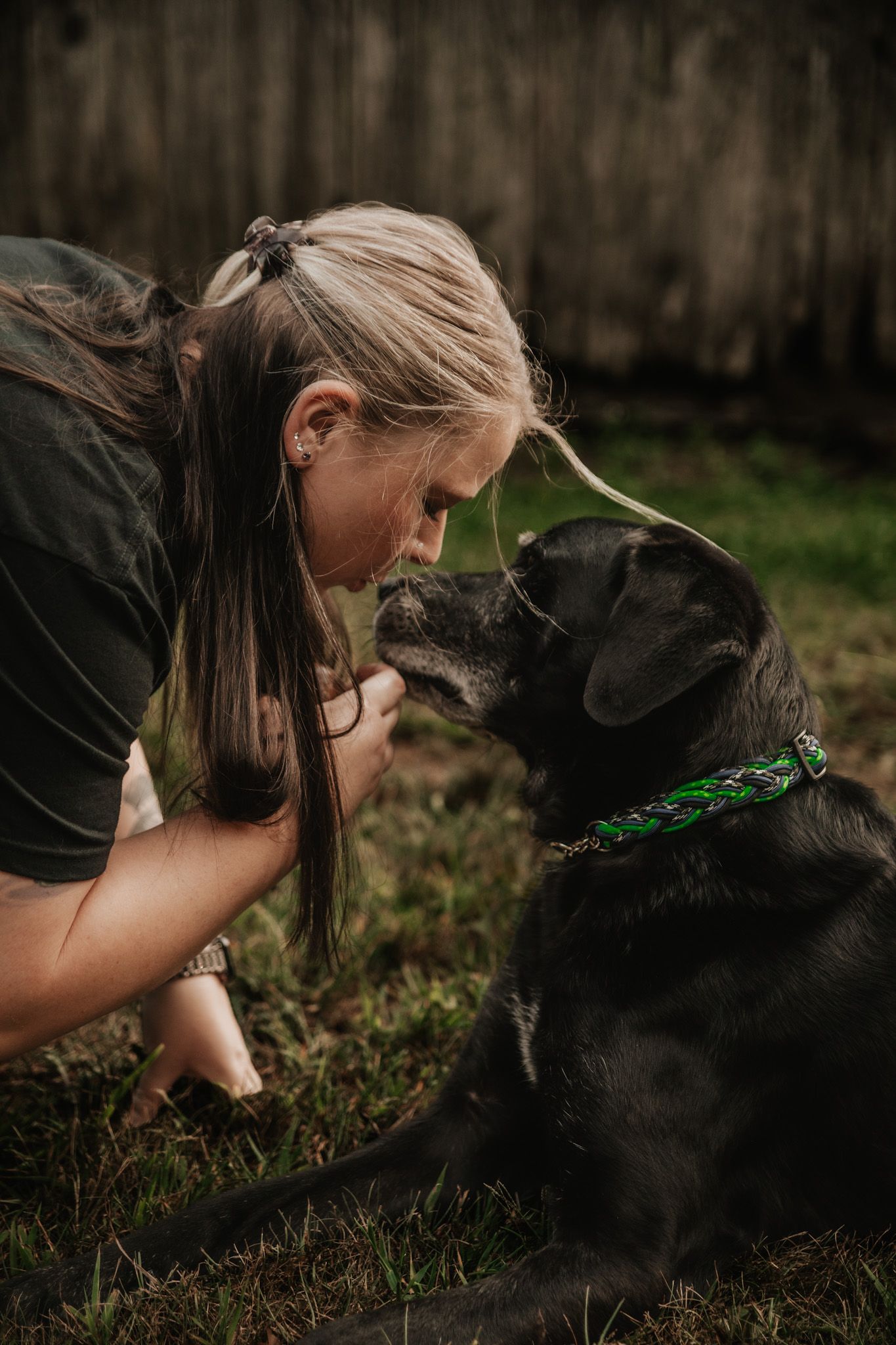A young blonde woman shares a tender nose-to-nose moment with a black Labrador wearing a green collar outdoors.