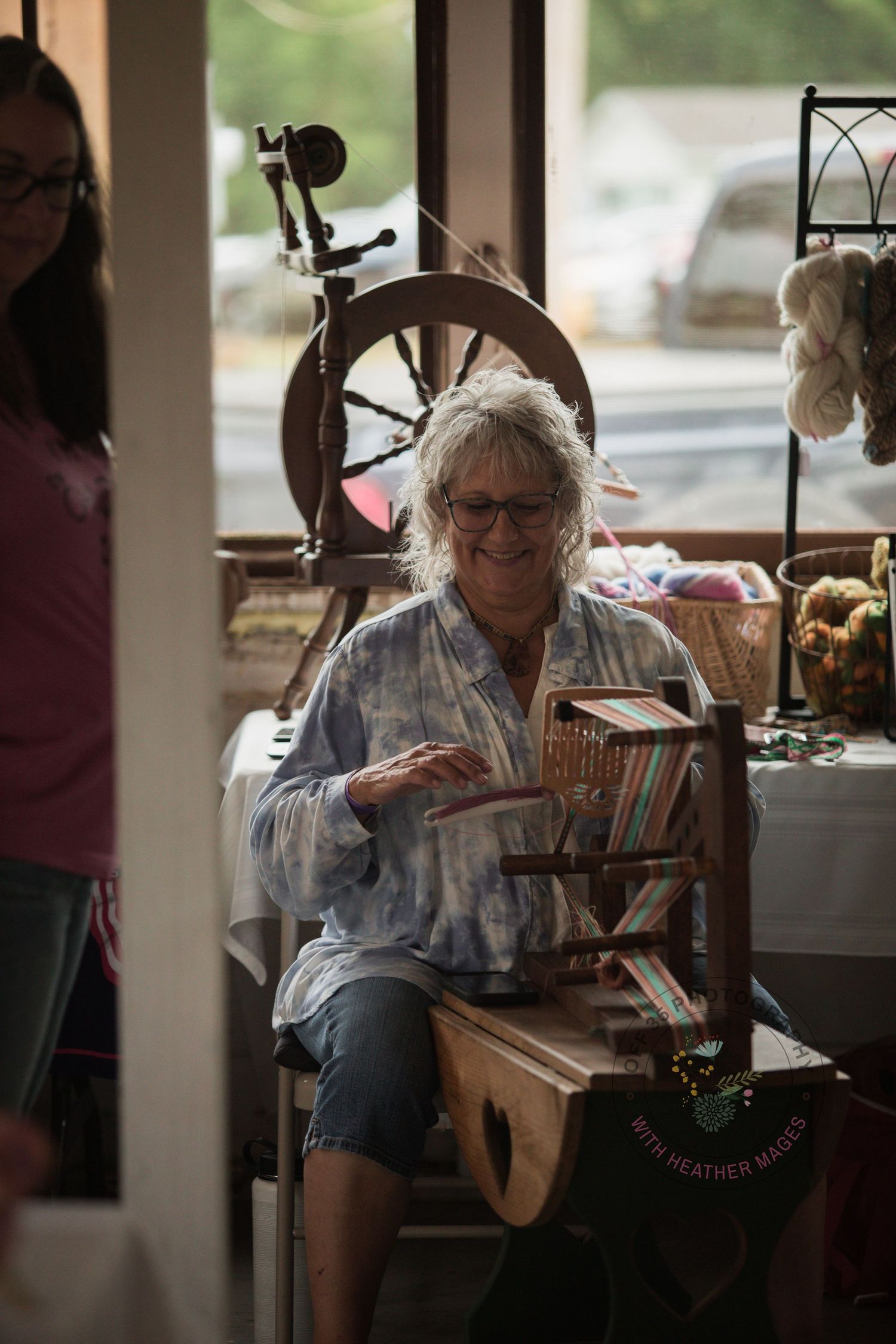 Someone sits at a spinning wheel in a cozy indoor setting with natural light streaming through windows.