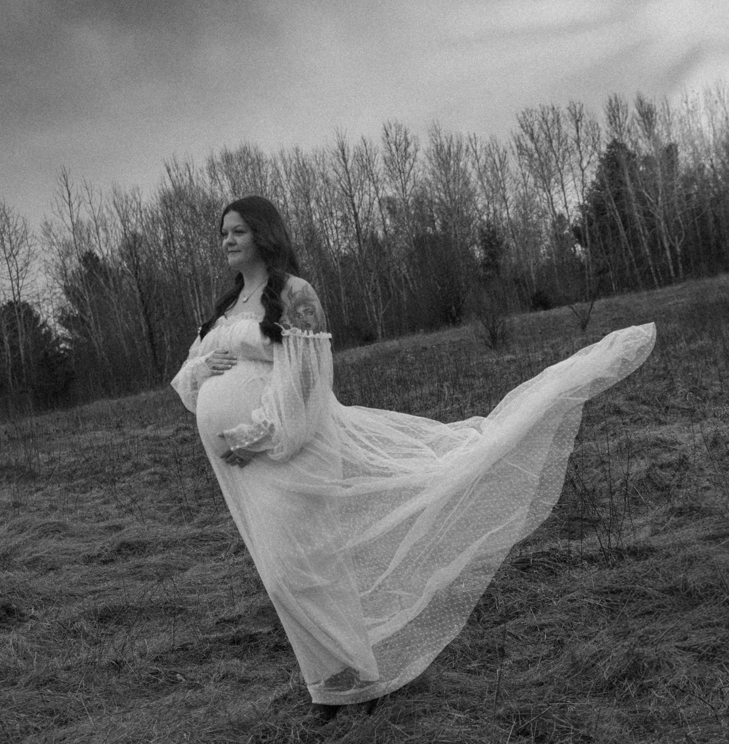 Black and white maternity photo of pregnant woman in flowing white dress twirling in open field.