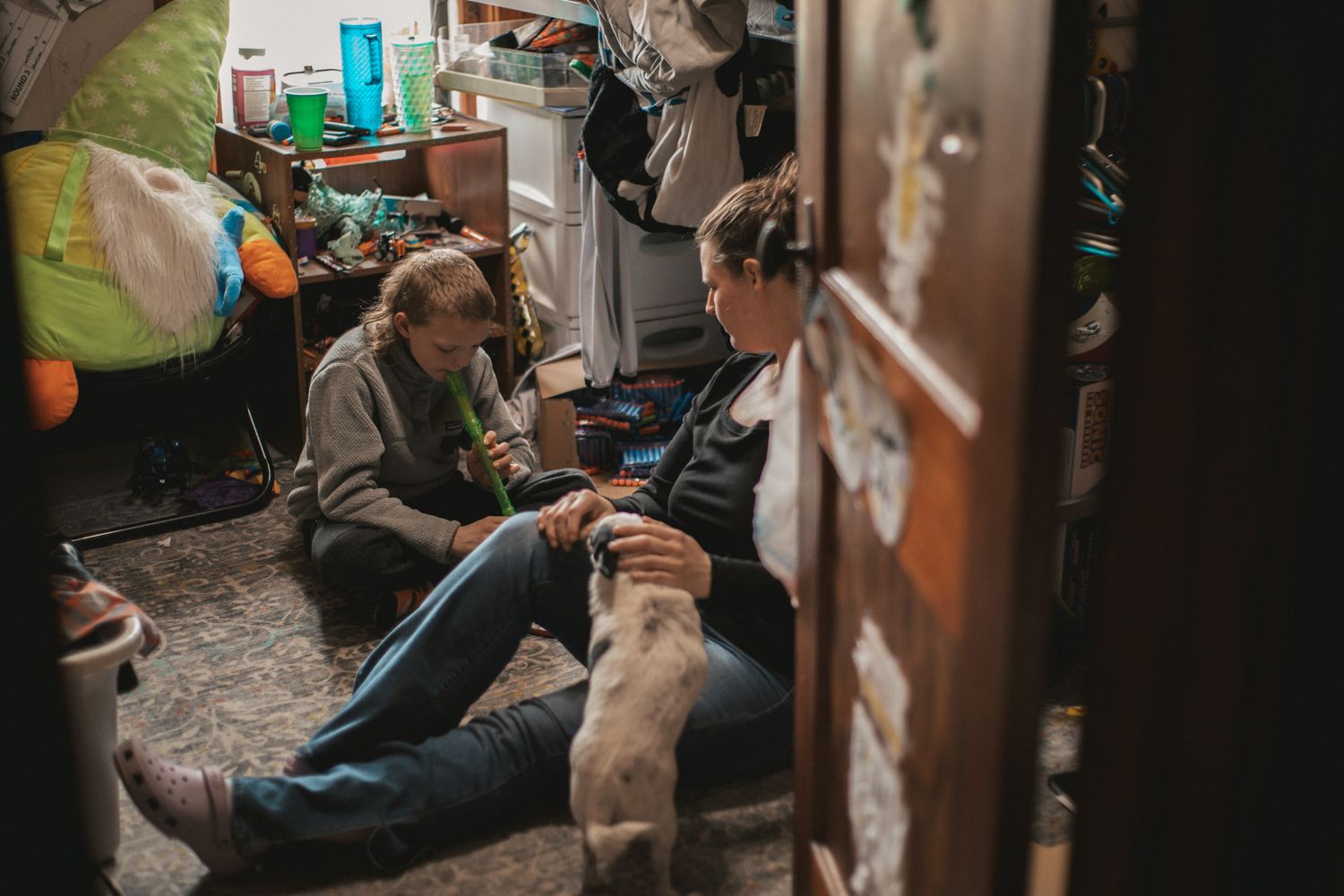 Two people sit on a cluttered floor petting a dog in a dimly lit, messy room.