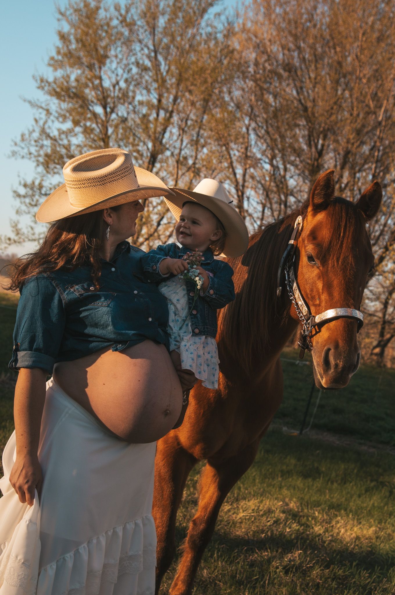 Pregnant woman in cowboy hat holds toddler near chestnut horse outdoors at golden hour.