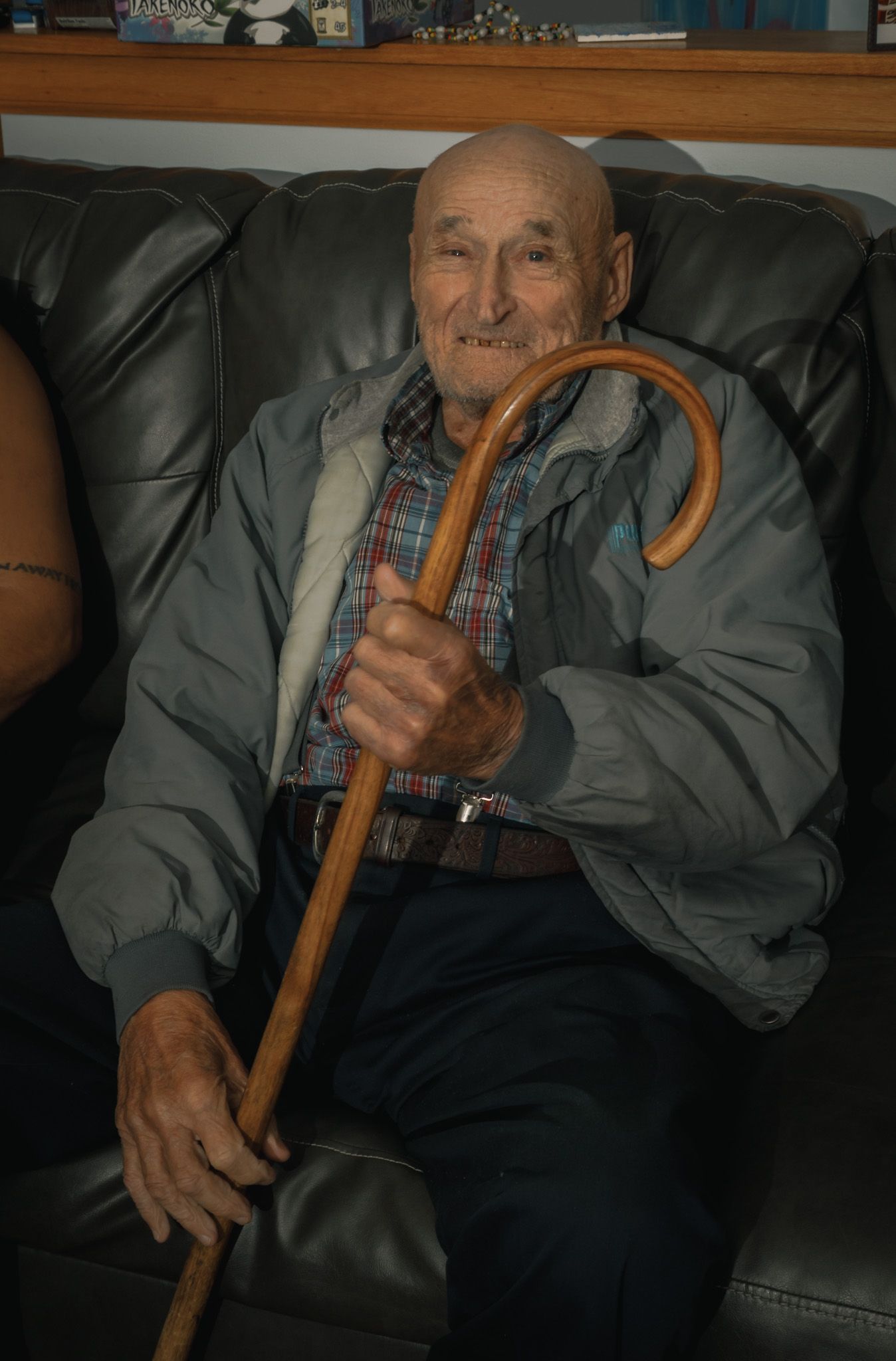 Smiling elderly bald man holds wooden cane while seated on leather sofa indoors.