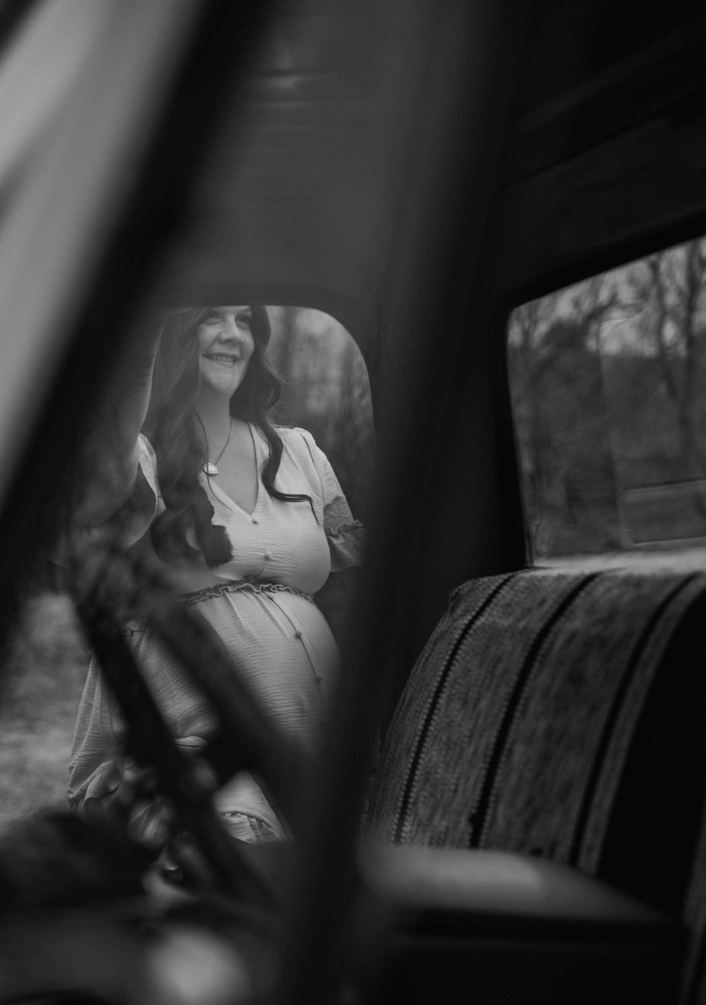 Black and white photo of a smiling pregnant woman in a vintage car, viewed through the window.