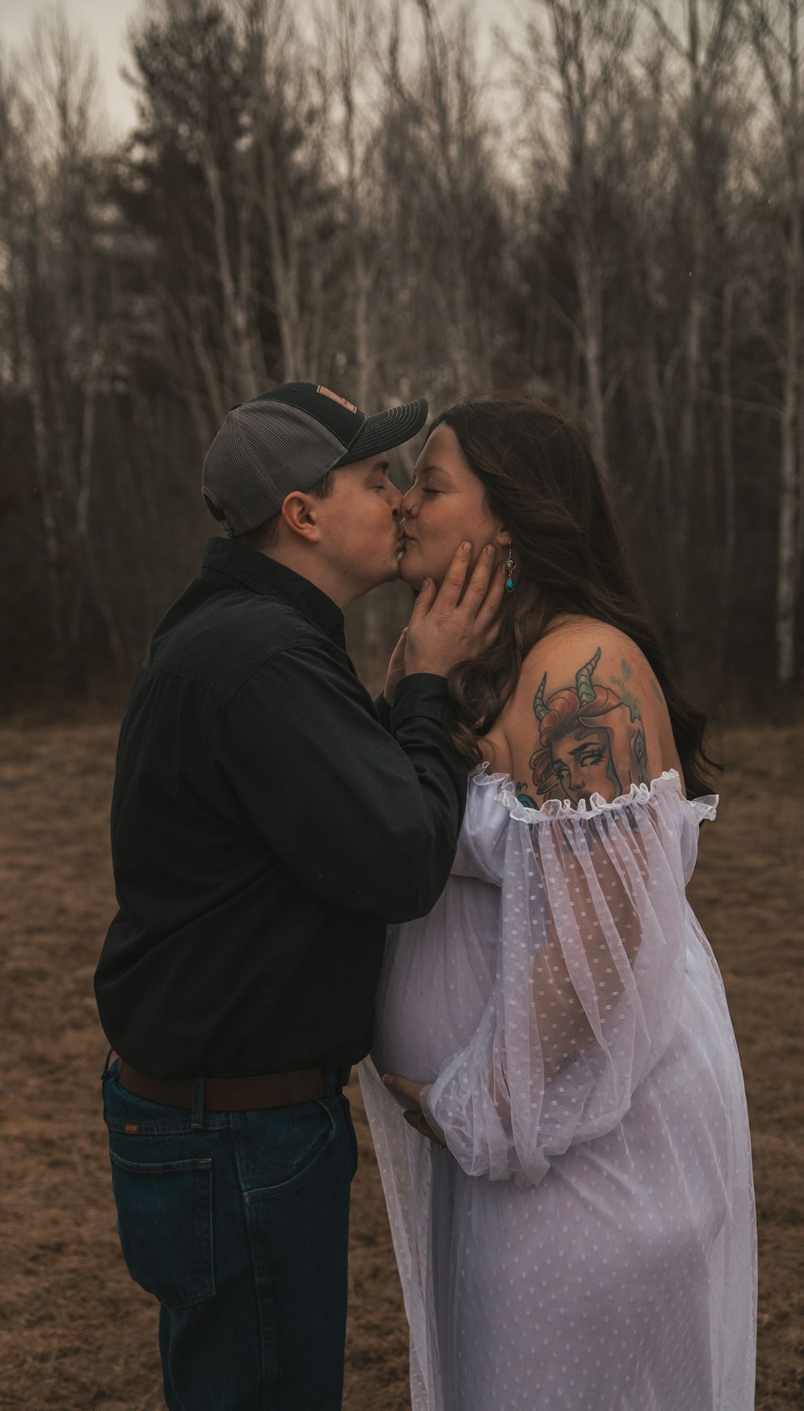 Couple sharing a romantic kiss outdoors during maternity photo shoot, woman in white dress with shoulder tattoo.