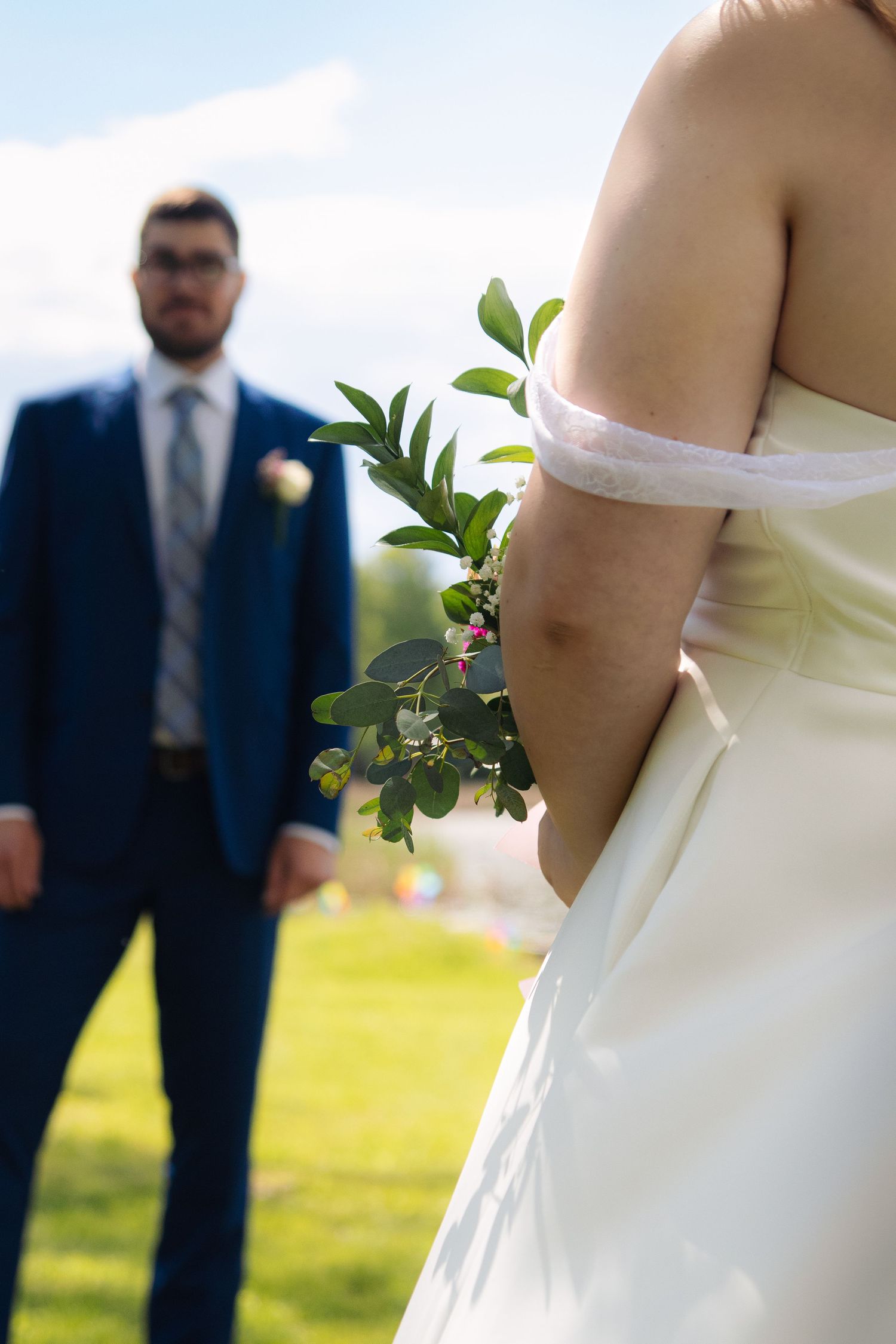 Close-up detail of a wedding dress and bouquet with a figure in a blue suit in the background by a lake.