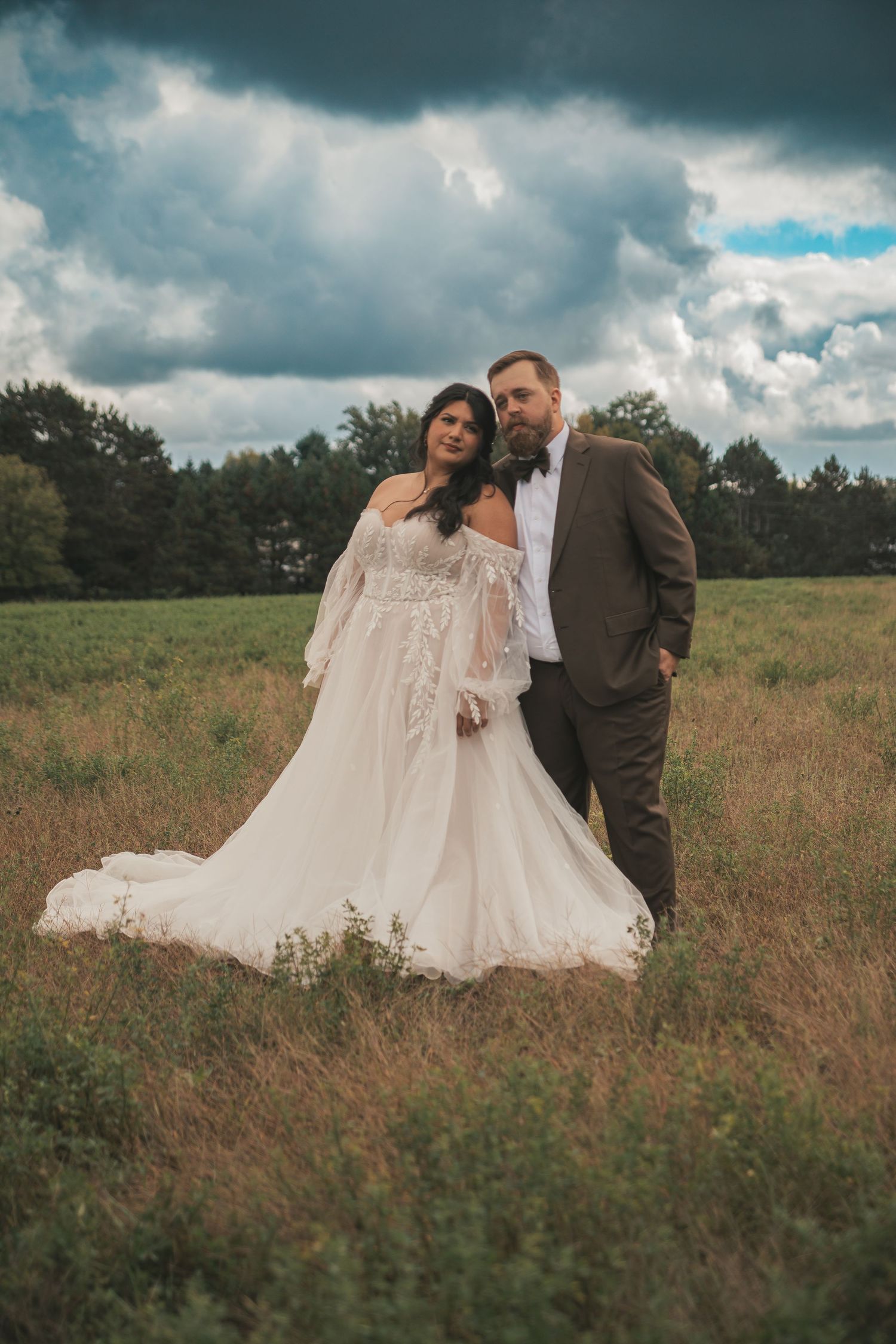 A couple poses together in a rustic meadow field with dramatic cloudy skies above during their outdoor photoshoot.