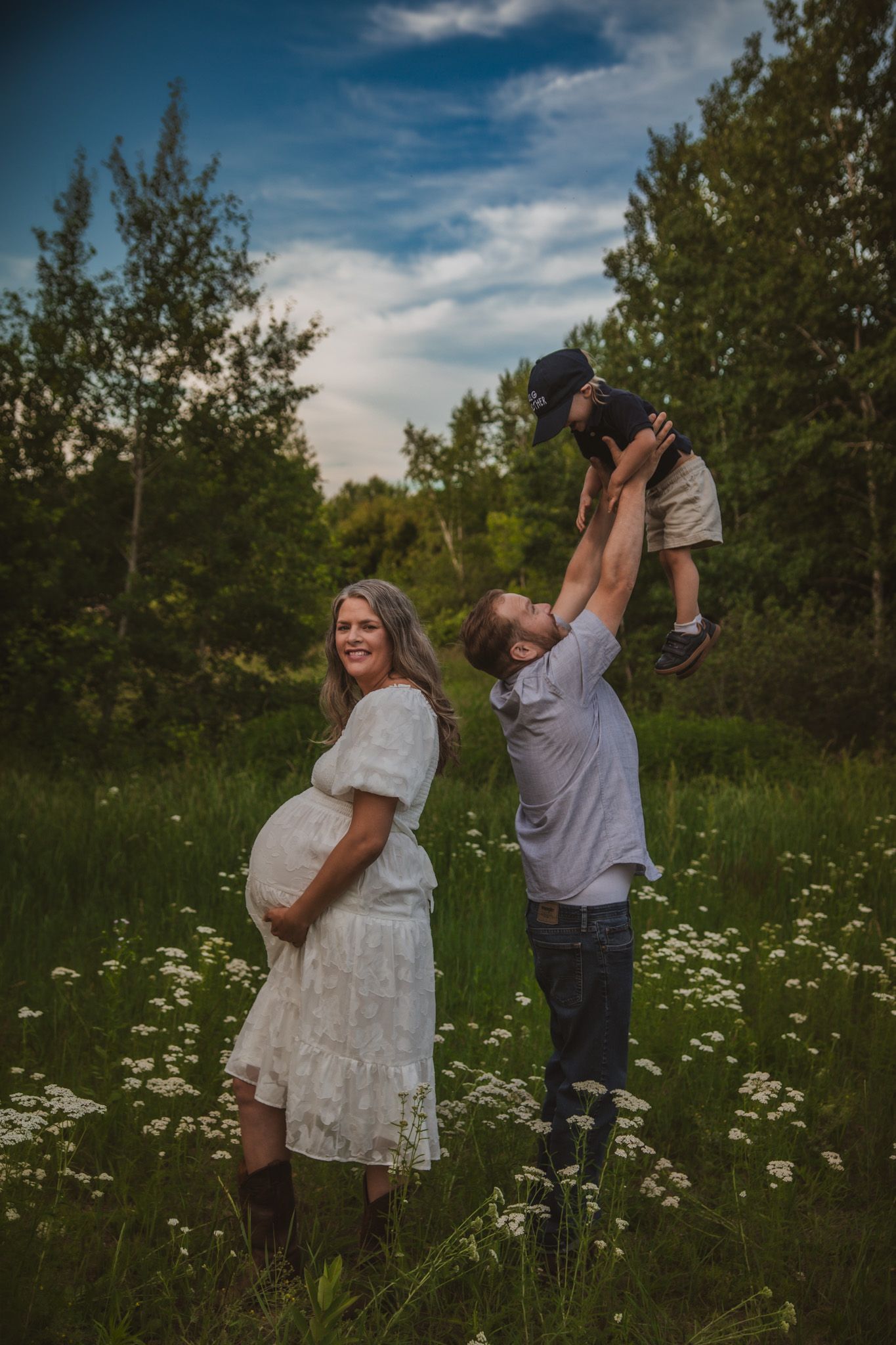 Pregnant woman in white dress stands beside man lifting toddler in sunny wildflower meadow surrounded by trees.
