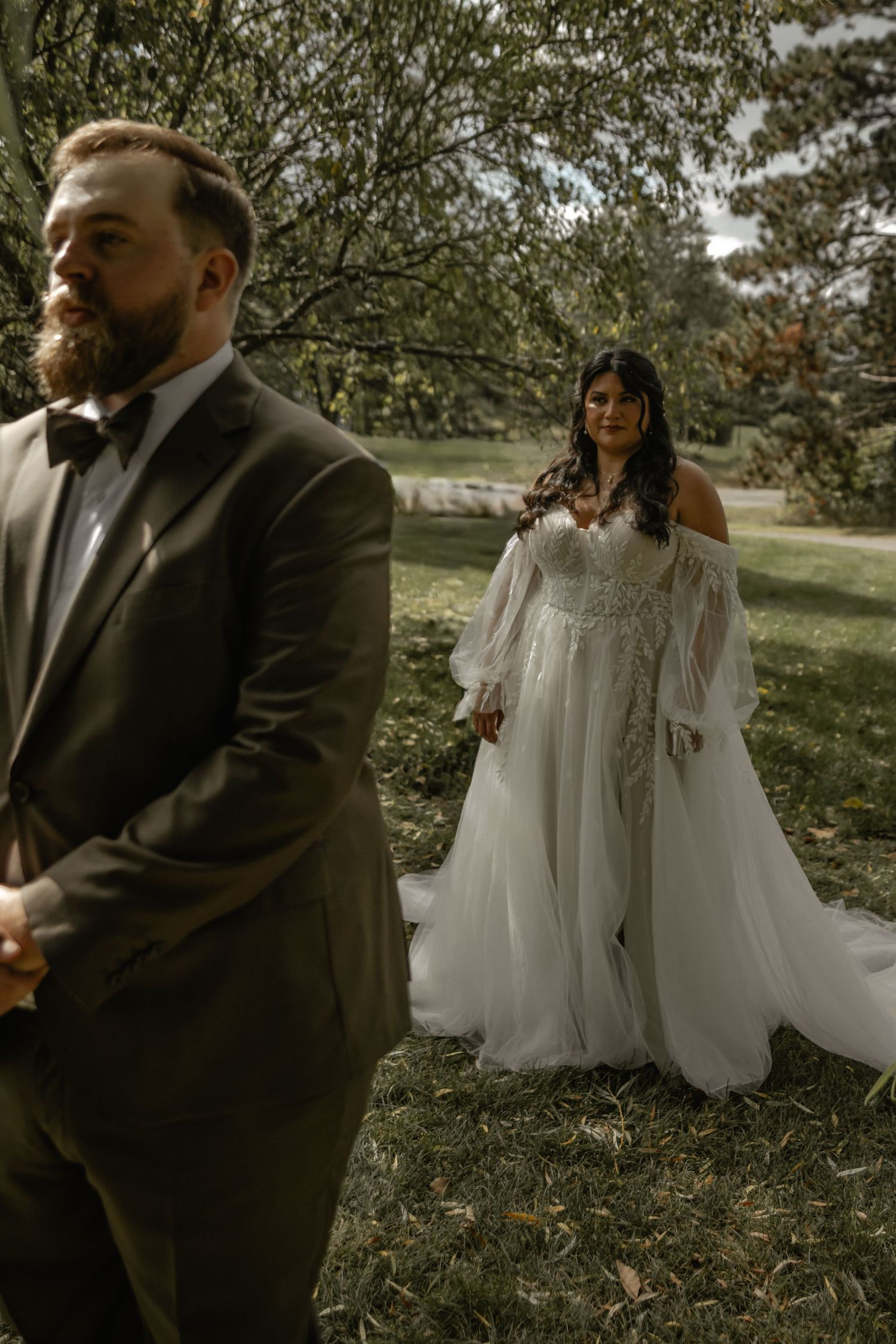 Bride in off-shoulder tulle gown stands behind bearded groom in gray suit with bow tie during outdoor wedding ceremony.