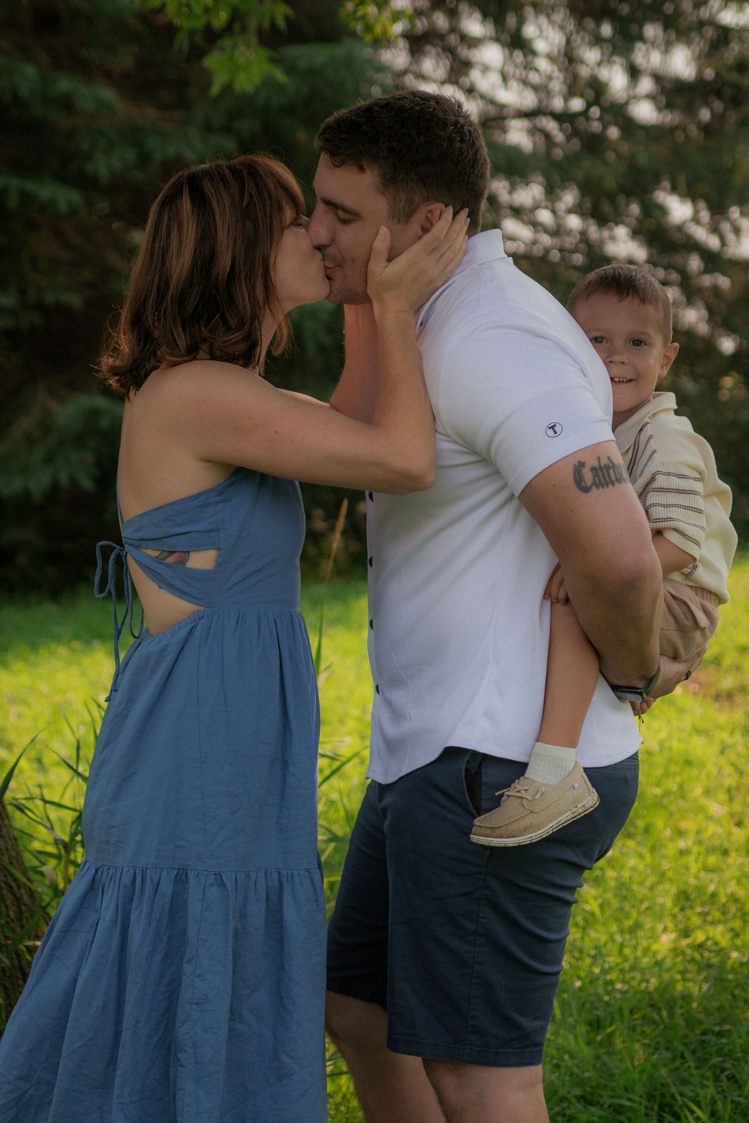 A couple shares a tender moment while holding a young child on a sunny day in a grassy outdoor setting.