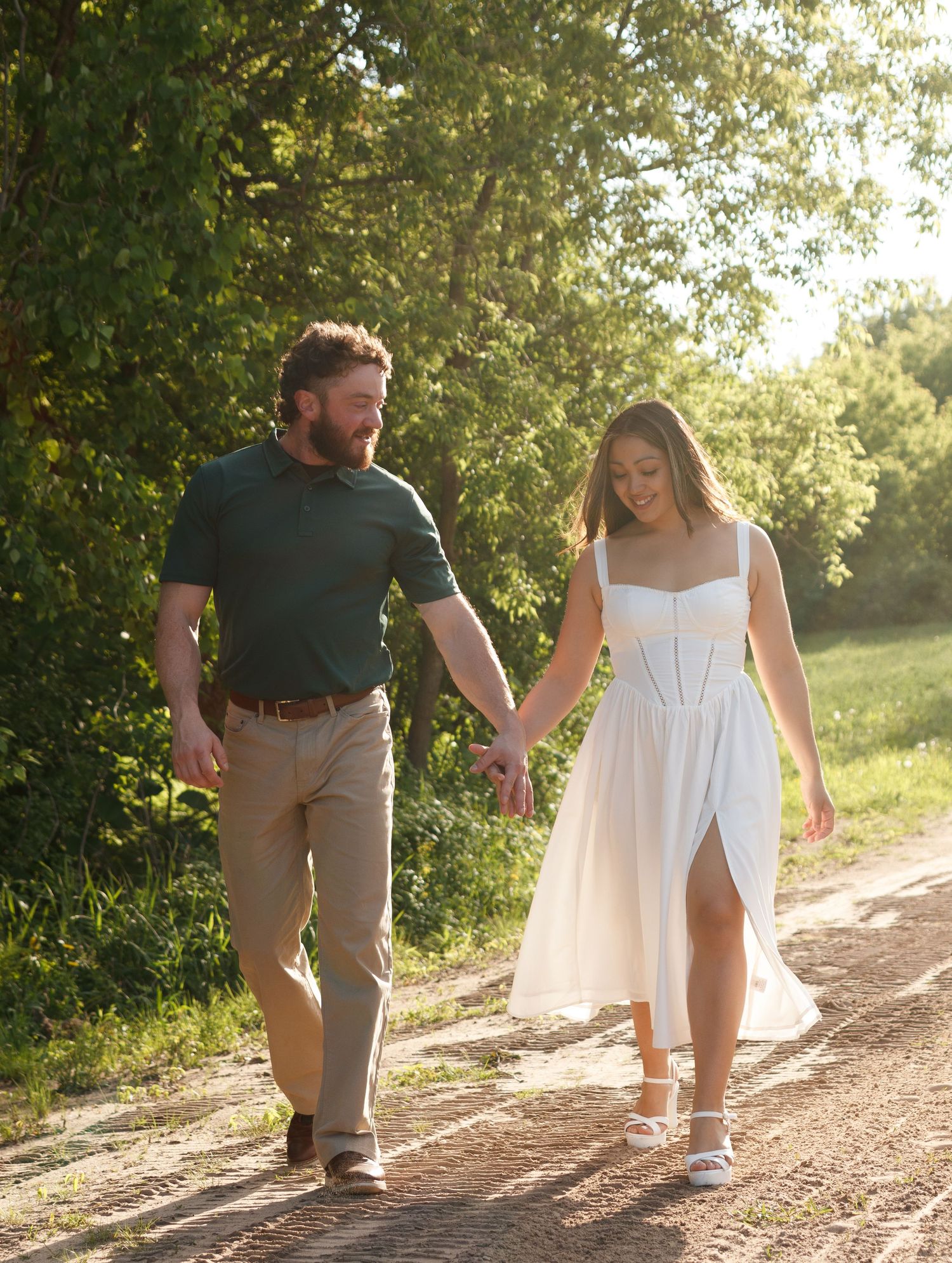 A couple walks hand in hand along a sunlit dirt path with green foliage in the background on a summer day.