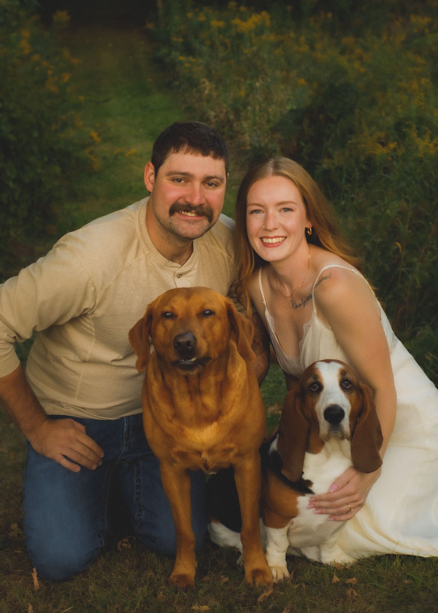 Smiling couple kneeling outdoors with a golden red Labrador and a Basset Hound in a lush green garden setting.